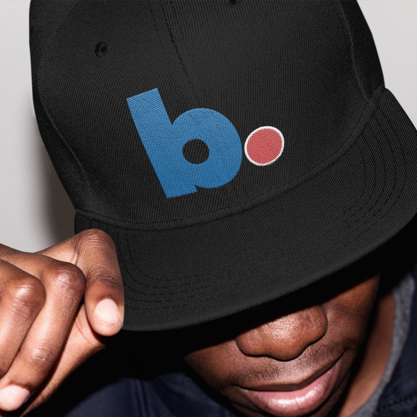 Black cap with a blue and red b. logo worn by a person.