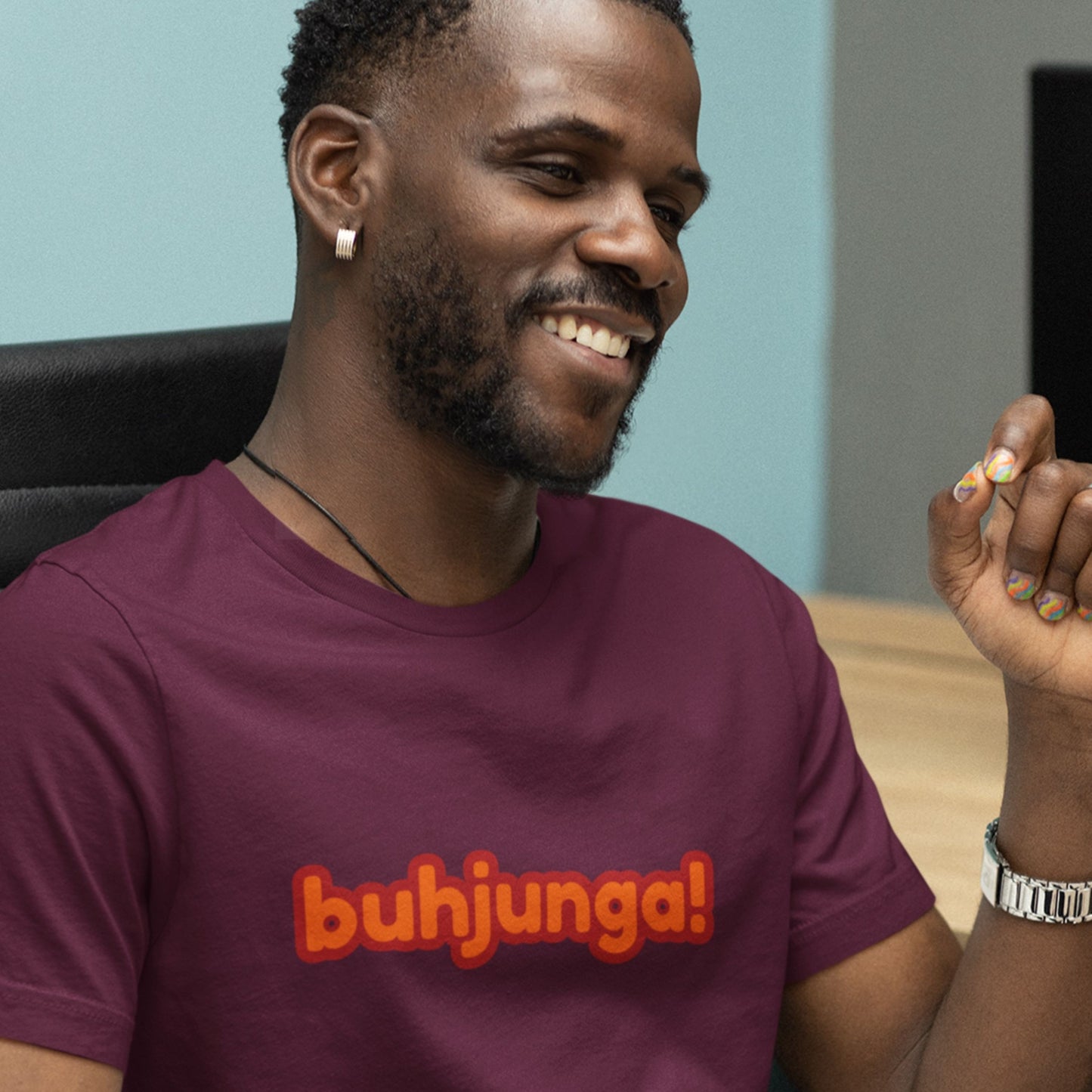 Seated man wearing a maroon t-shirt with multicolored 'buhjunga!' text in an office.