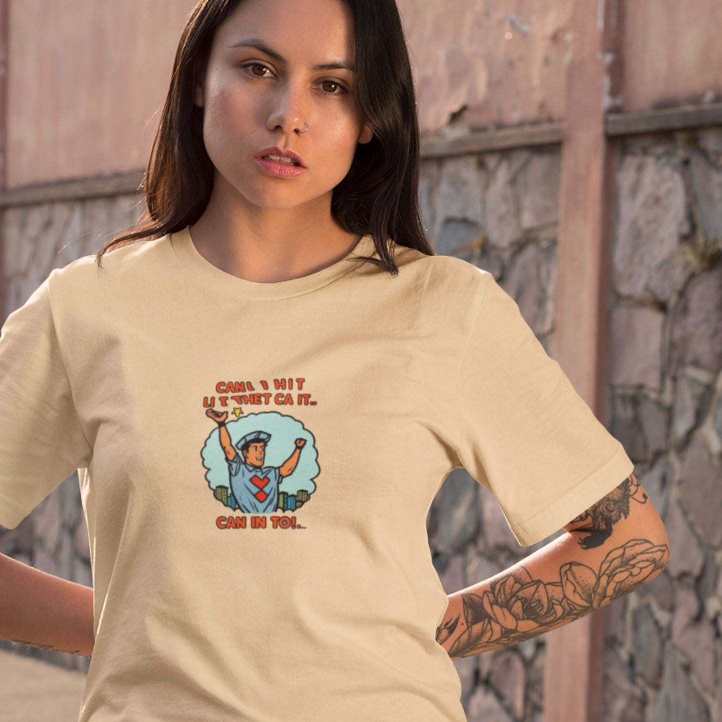 Woman wearing a beige t-shirt with a graphic design and text, standing against a stone wall.