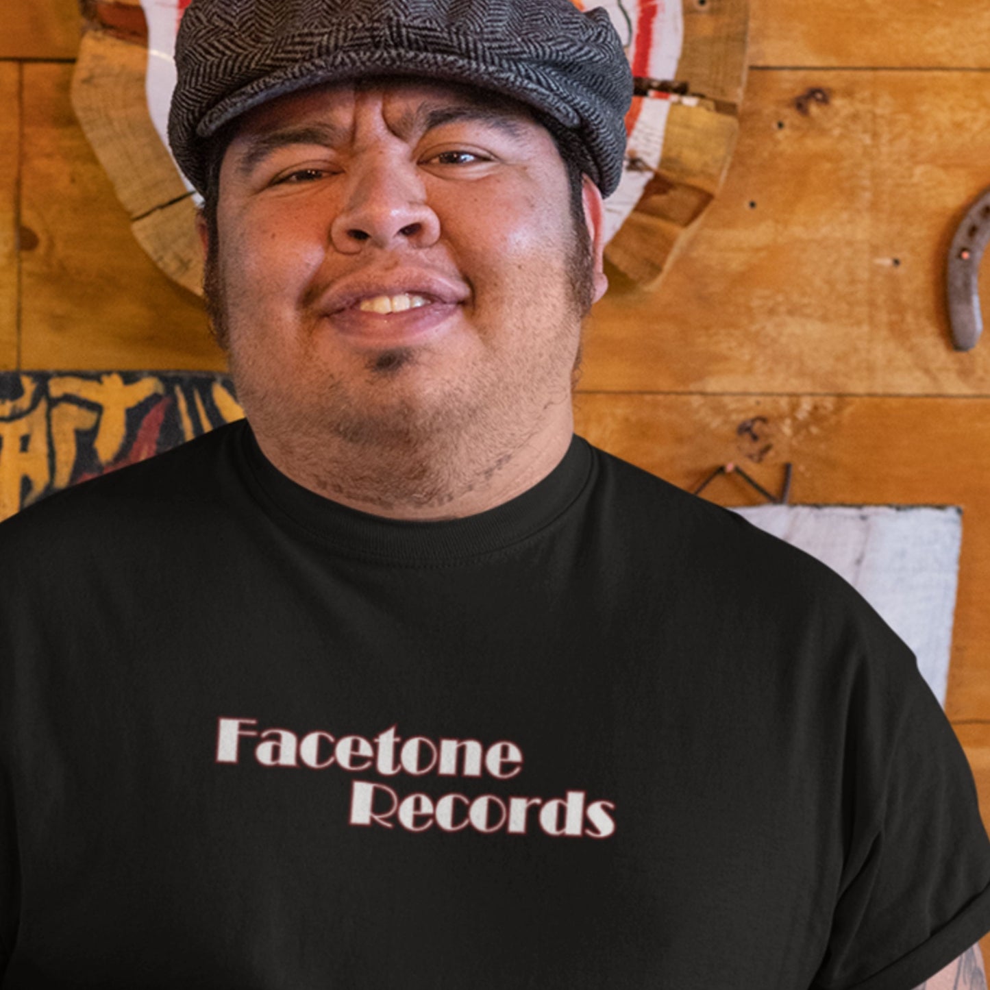 Man wearing a black 'Facetone Records' t-shirt in front of a wooden wall with decorative items.
