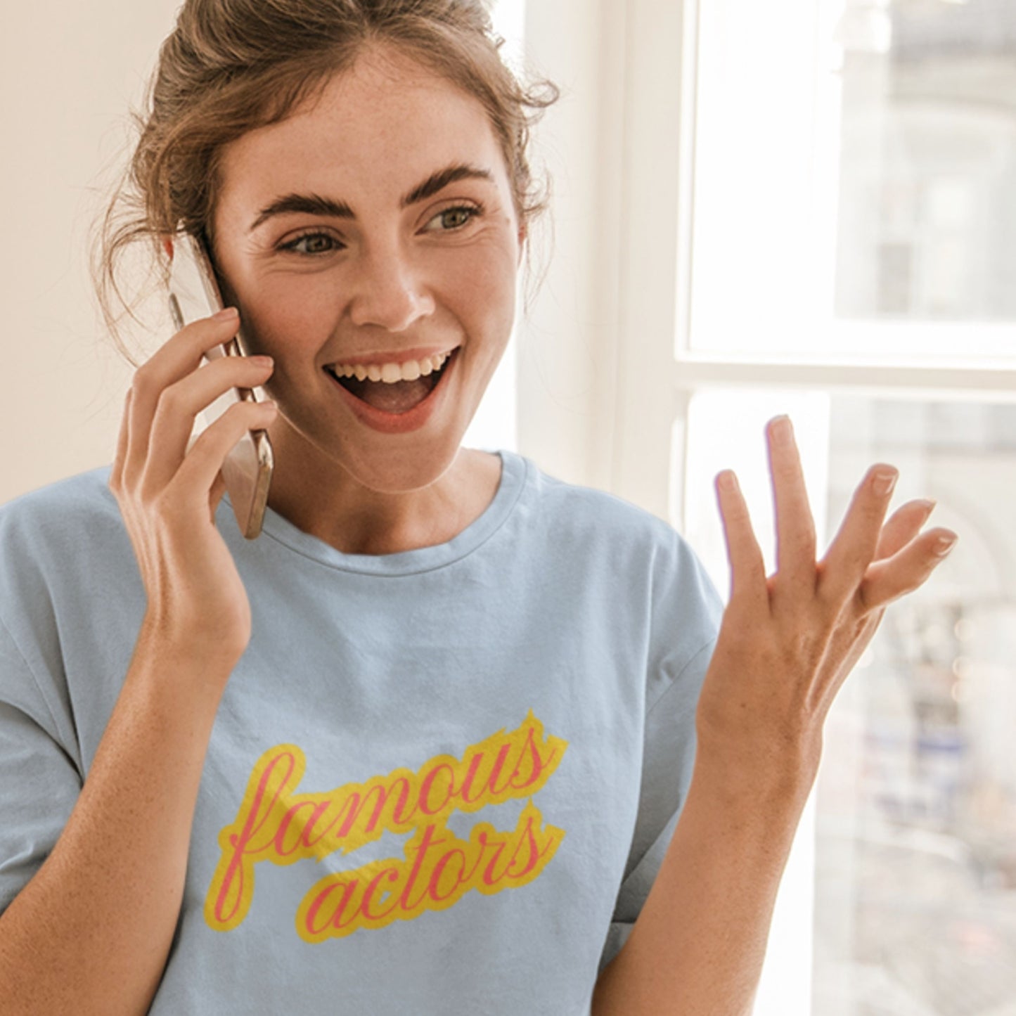 Happy woman wearing a baby blue t-shirt with multicolored 'famous actors' text talking on a phone in a room.