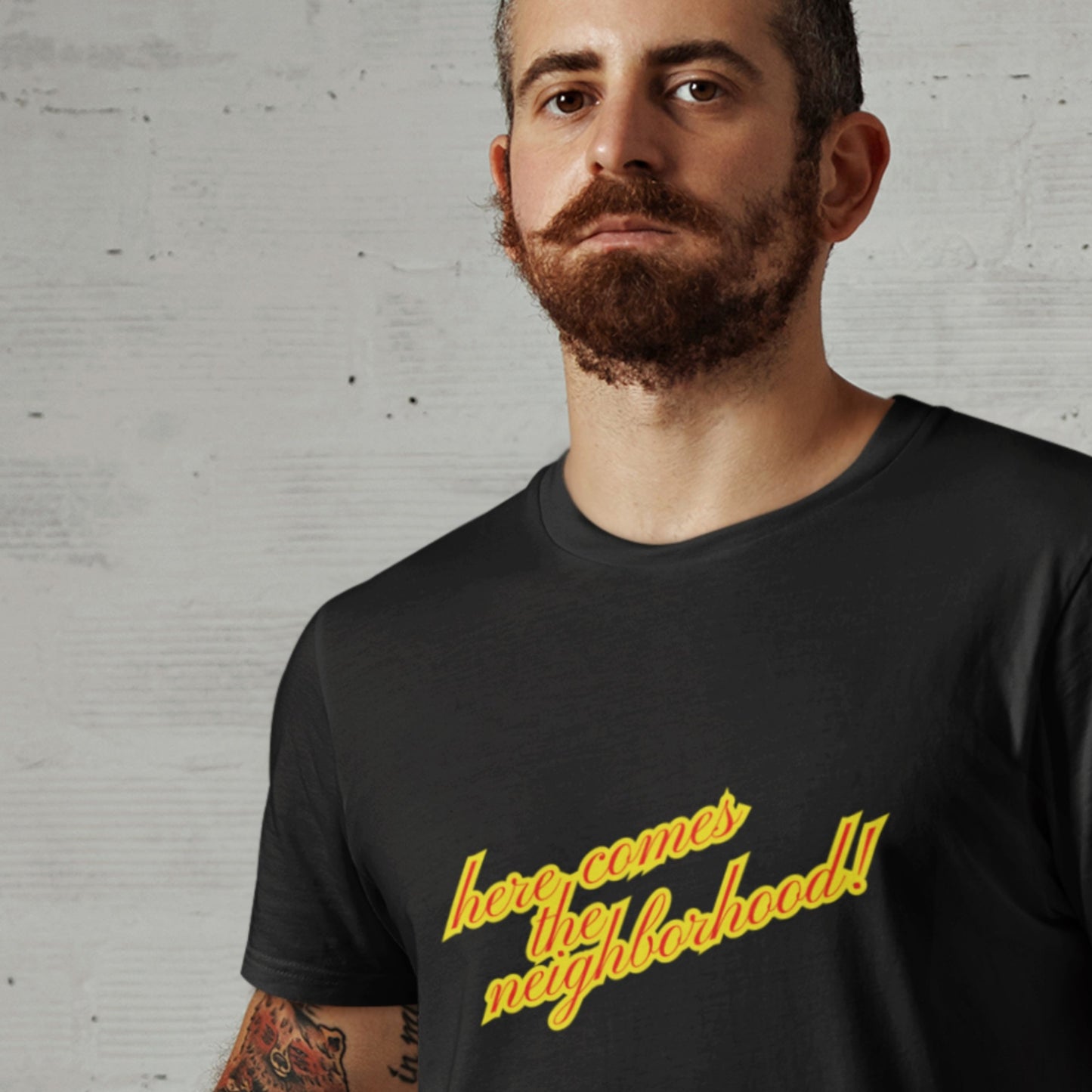 Apprehensive man wearing a black t-shirt with multicolored 'Here Comes the Neighborhood!' text on a grey background.