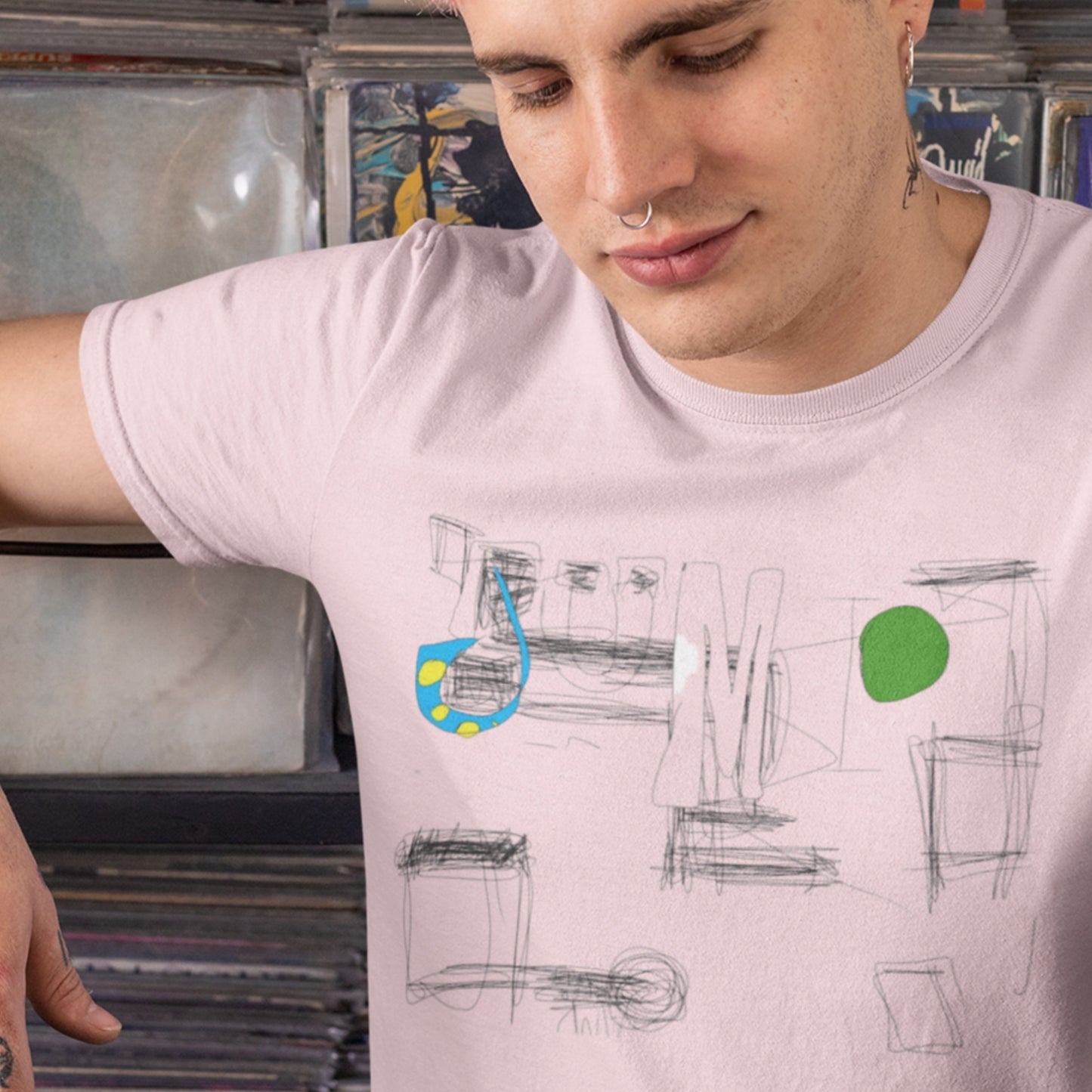 Person wearing a light pink t-shirt with abstract designs and colorful elements, standing in front of a stack of records.