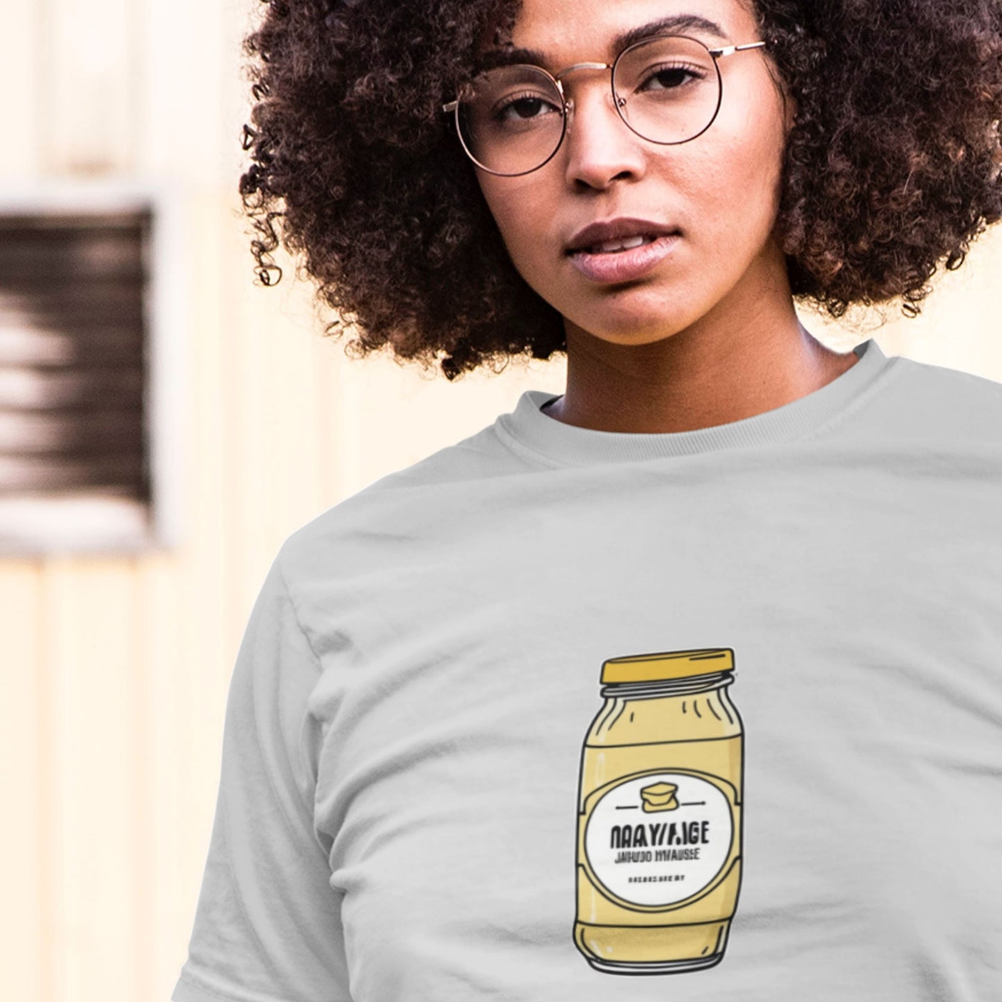 Woman wearing a light grey shirt with a drawing of a jar of specialty Dijonaise against an outdoor beige background.