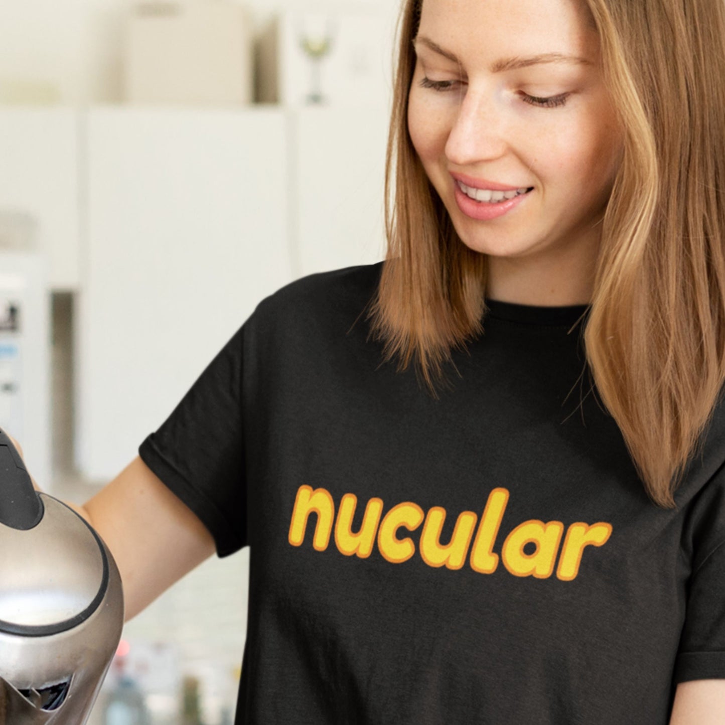 Woman wearing a black t-shirt with 'nucular' printed in yellow, holding a coffee maker.