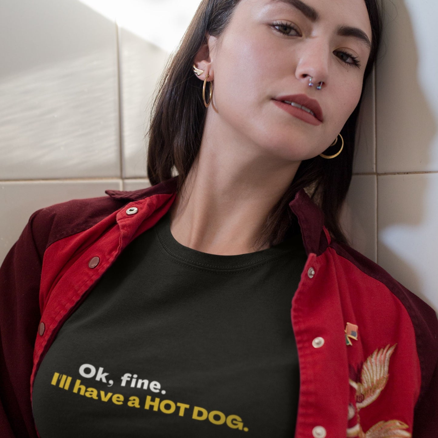 Woman wearing a black t-shirt with text and a red jacket in a tiled room.