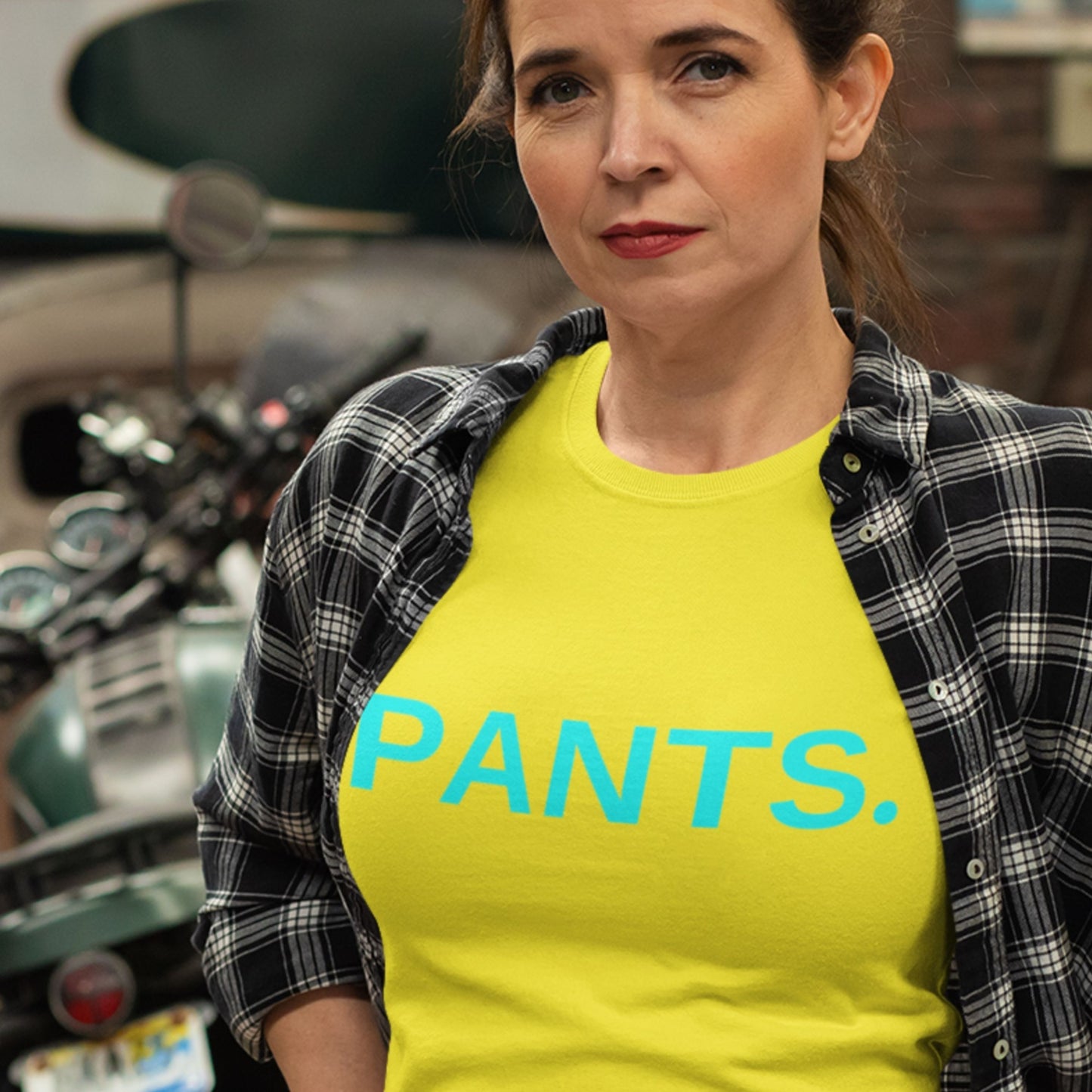 Woman wearing a yellow 'PANTS.' shirt with a plaid jacket, standing in front of a motorcycle.