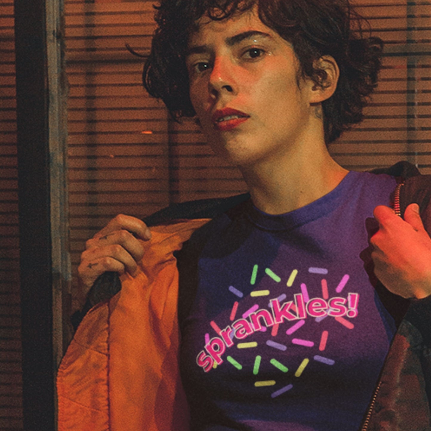 Person wearing a purple shirt with 'Pink Sprankles!' text, holding an orange jacket indoors.