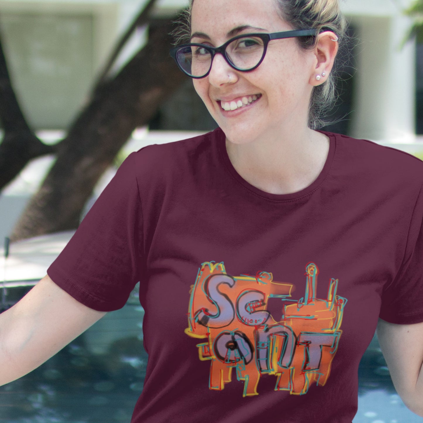 Woman in glasses wearing a maroon t-shirt with a colorful abstract drawing outdoors at a marina.