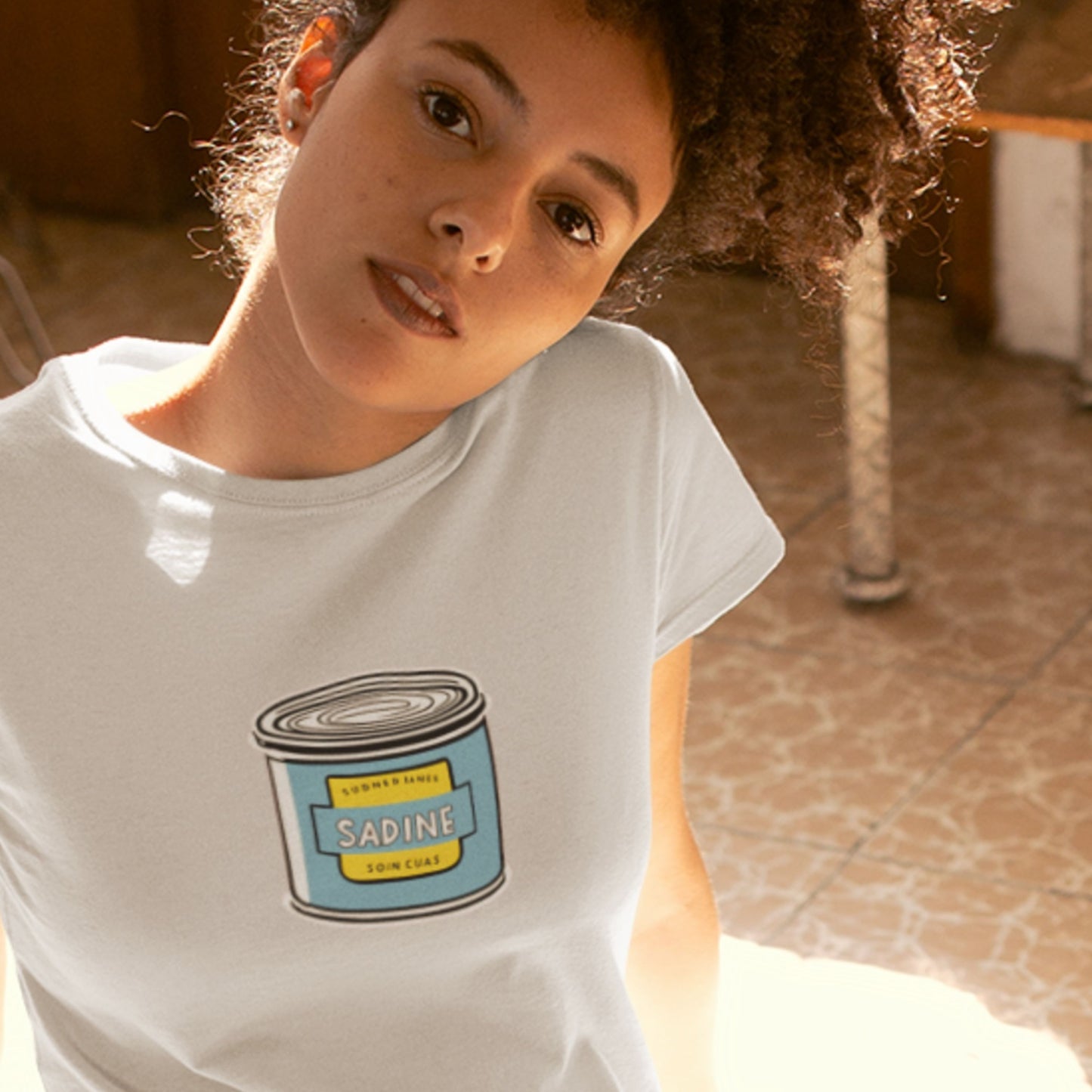 Woman wearing a light grey shirt with an illustration of a can of fish in a room.