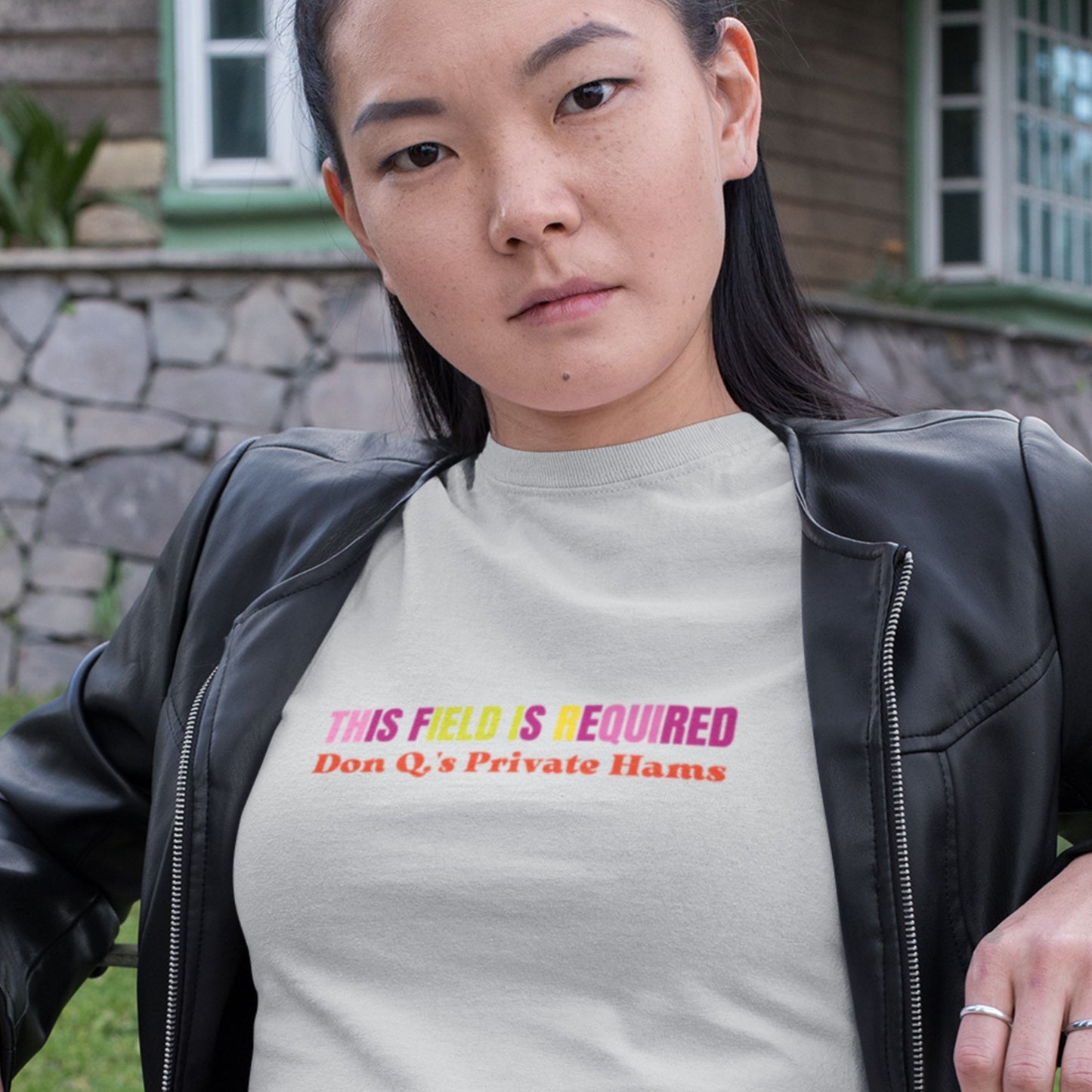 Woman wearing a light grey t-shirt with nonsensical, multicolored text leaning against a fence.