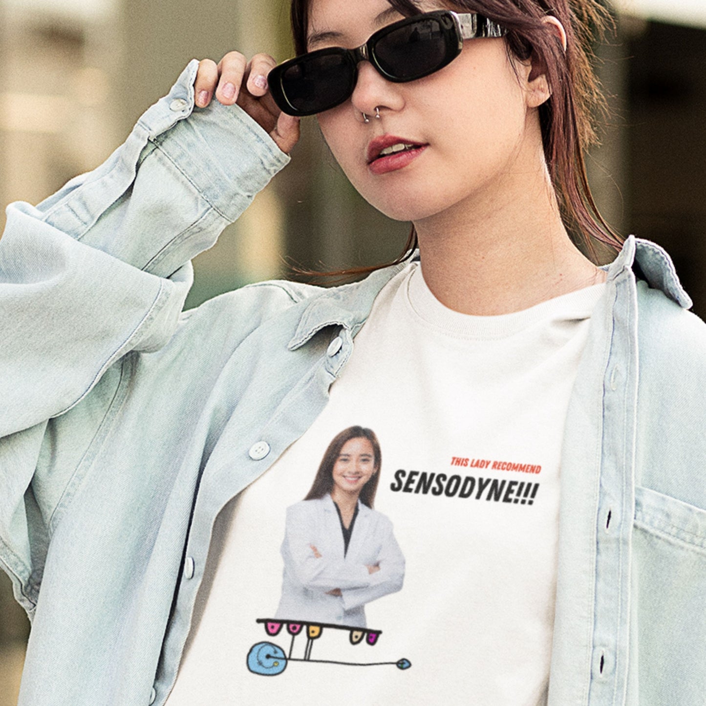 Woman in sunglasses wearing a white t-shirt with a colorful abstract drawing, humorous text and an image of a dentist outside.