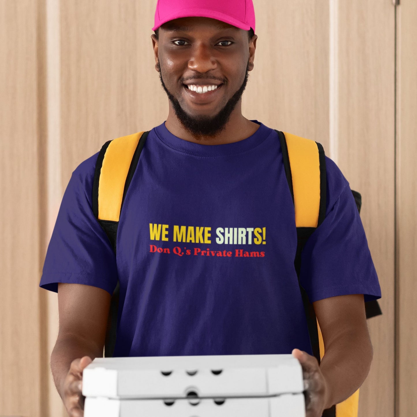 Man wearing a purple t-shirt with 'We Make Shirts!' text, holding a box against a beige curtain background.