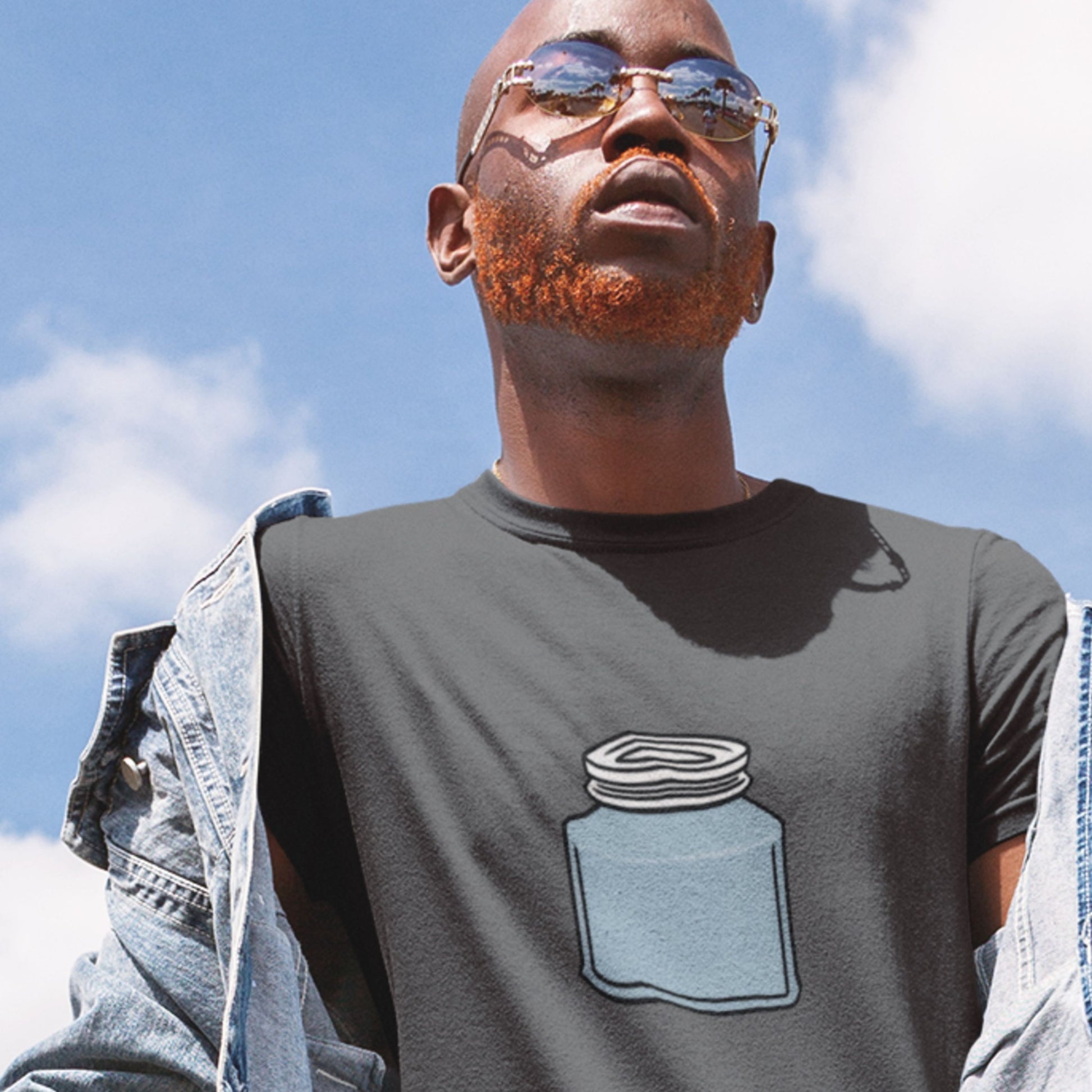Man wearing a grey shirt with a drawing of a blue jar with a screw top lid against a cloudy sky.