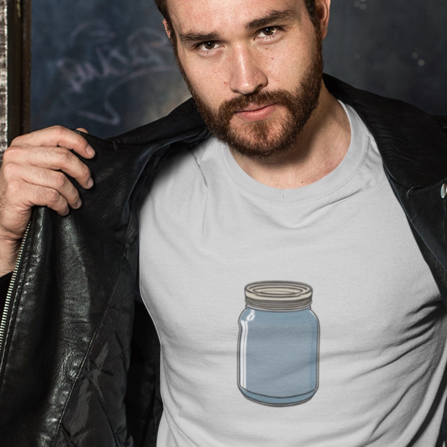 Man wearing a light grey t-shirt with a drawing of a blue mason jar in an interior space.