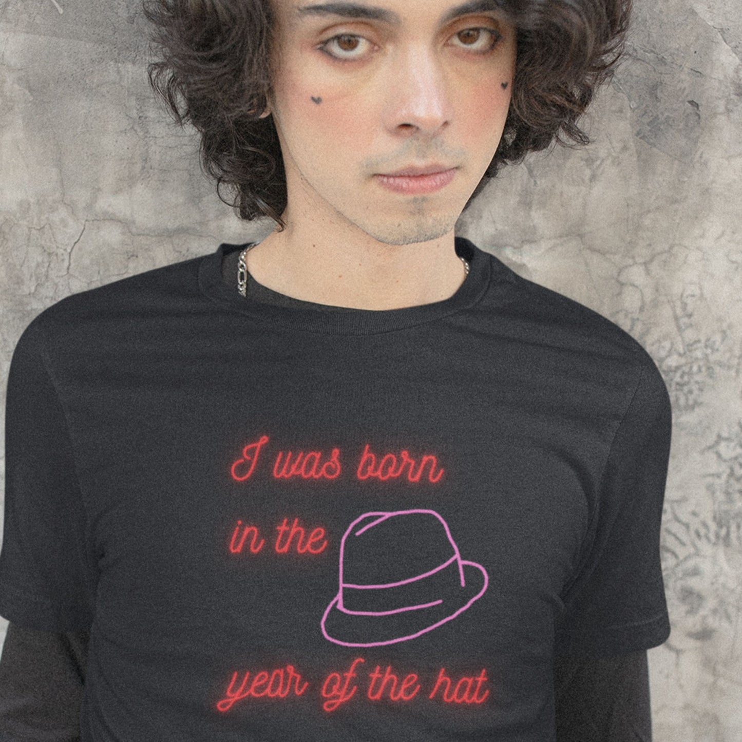 Person wearing a black t-shirt with red text and a pink hat graphic against a textured wall.