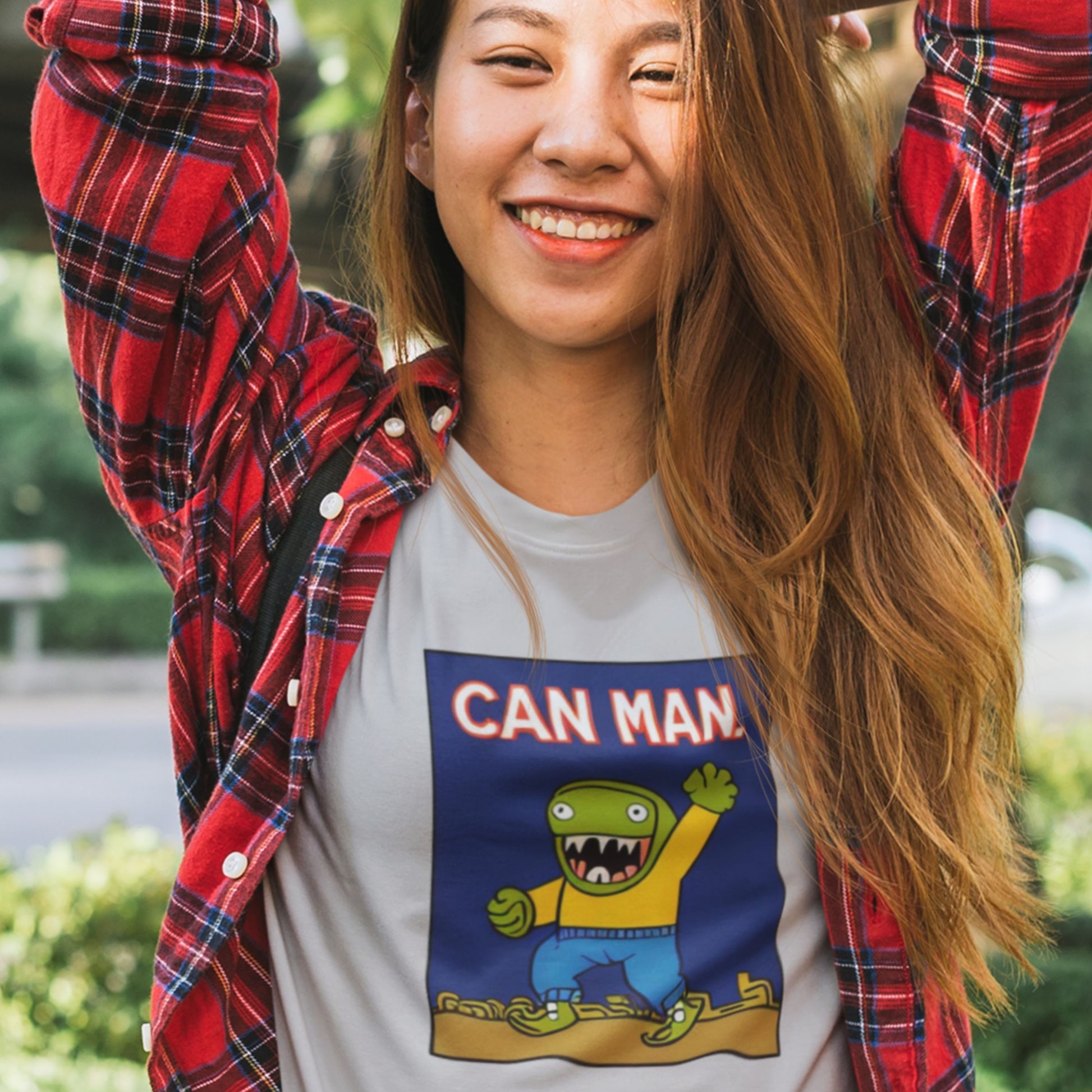 Person wearing a t-shirt with 'Can Man' graphic and red plaid shirt outdoors.