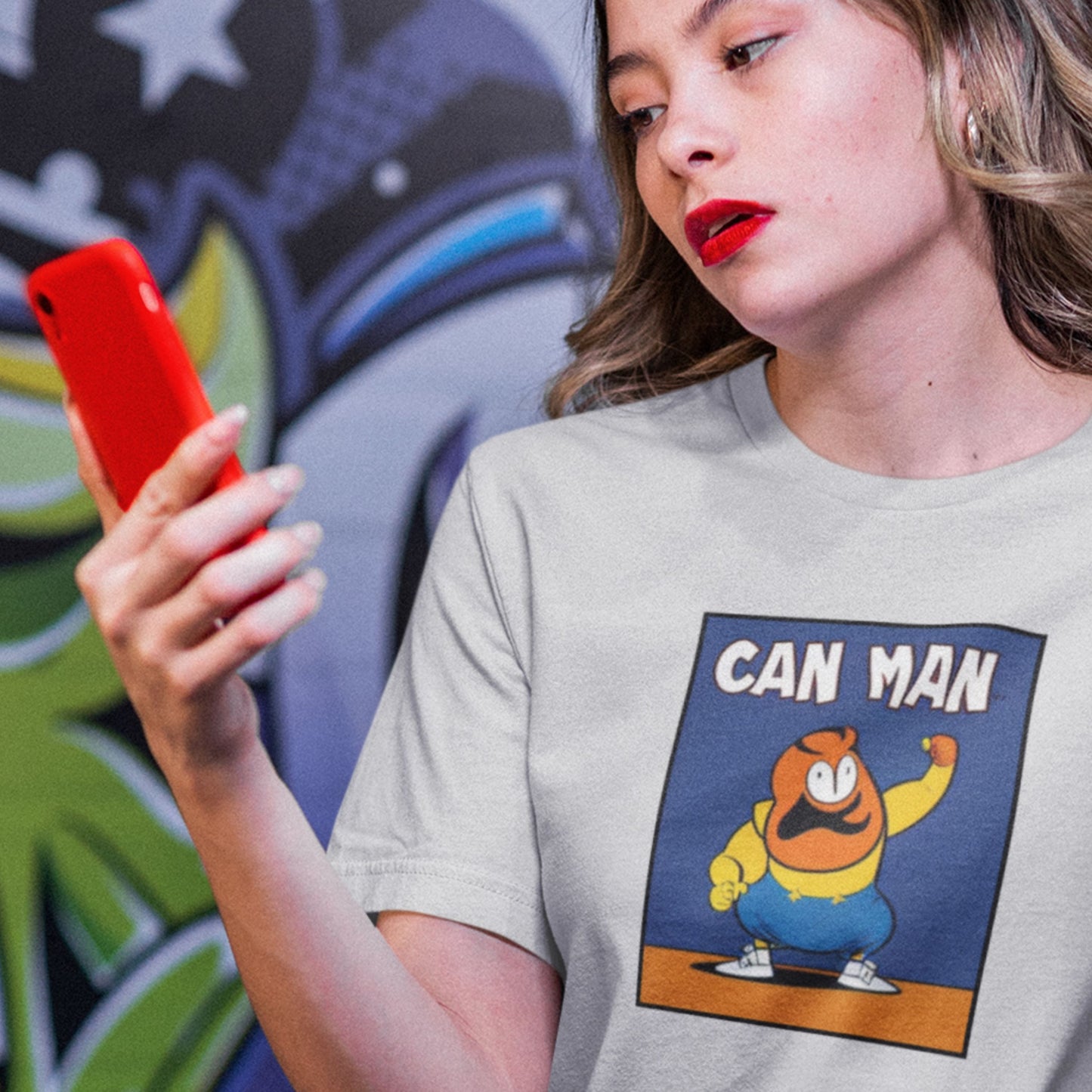 Person wearing a t-shirt with a cartoon character and 'CAN MAN' text, holding a red phone in front of a colorful graffiti wall.