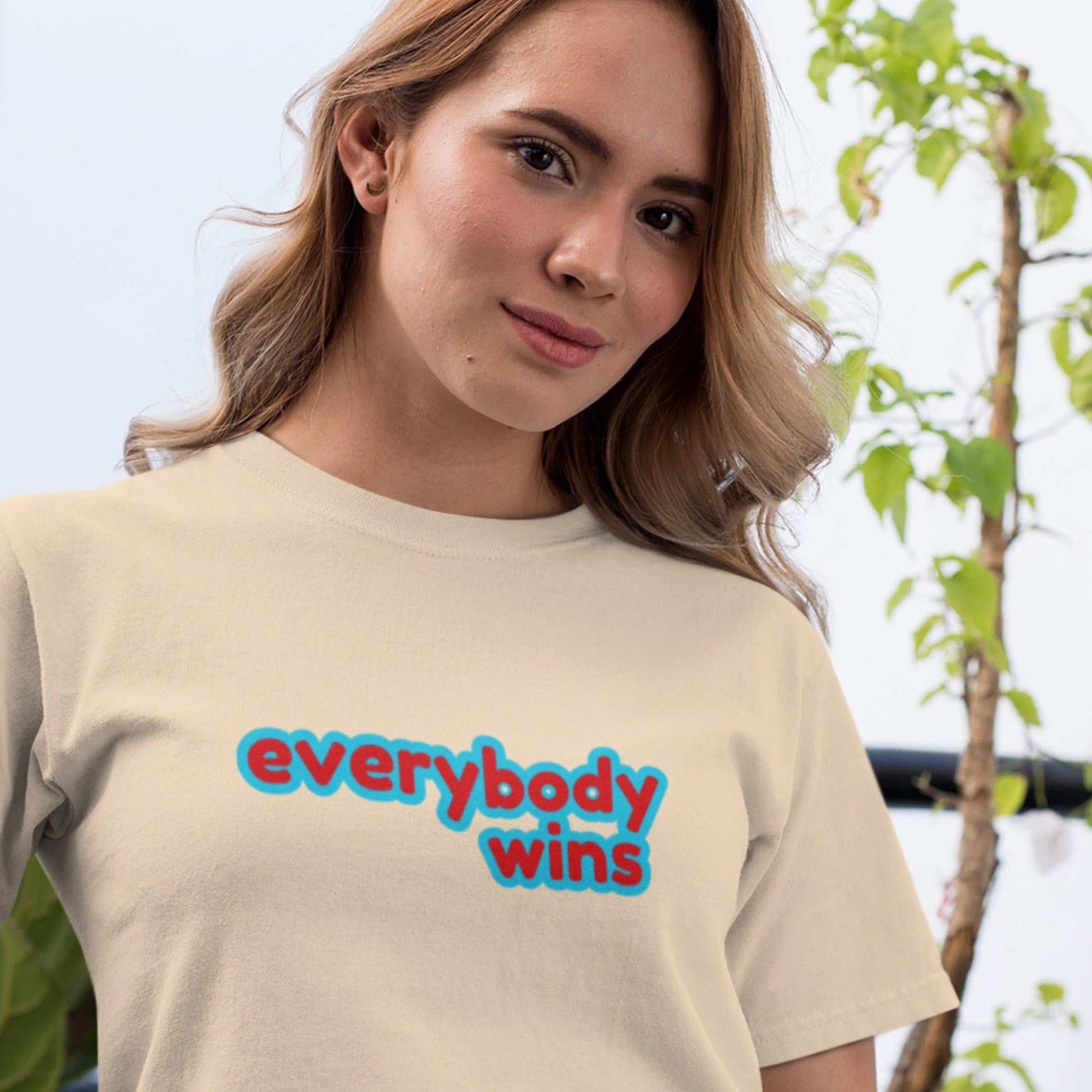 Woman wearing a beige t-shirt with 'everybody wins' text, standing outdoors.