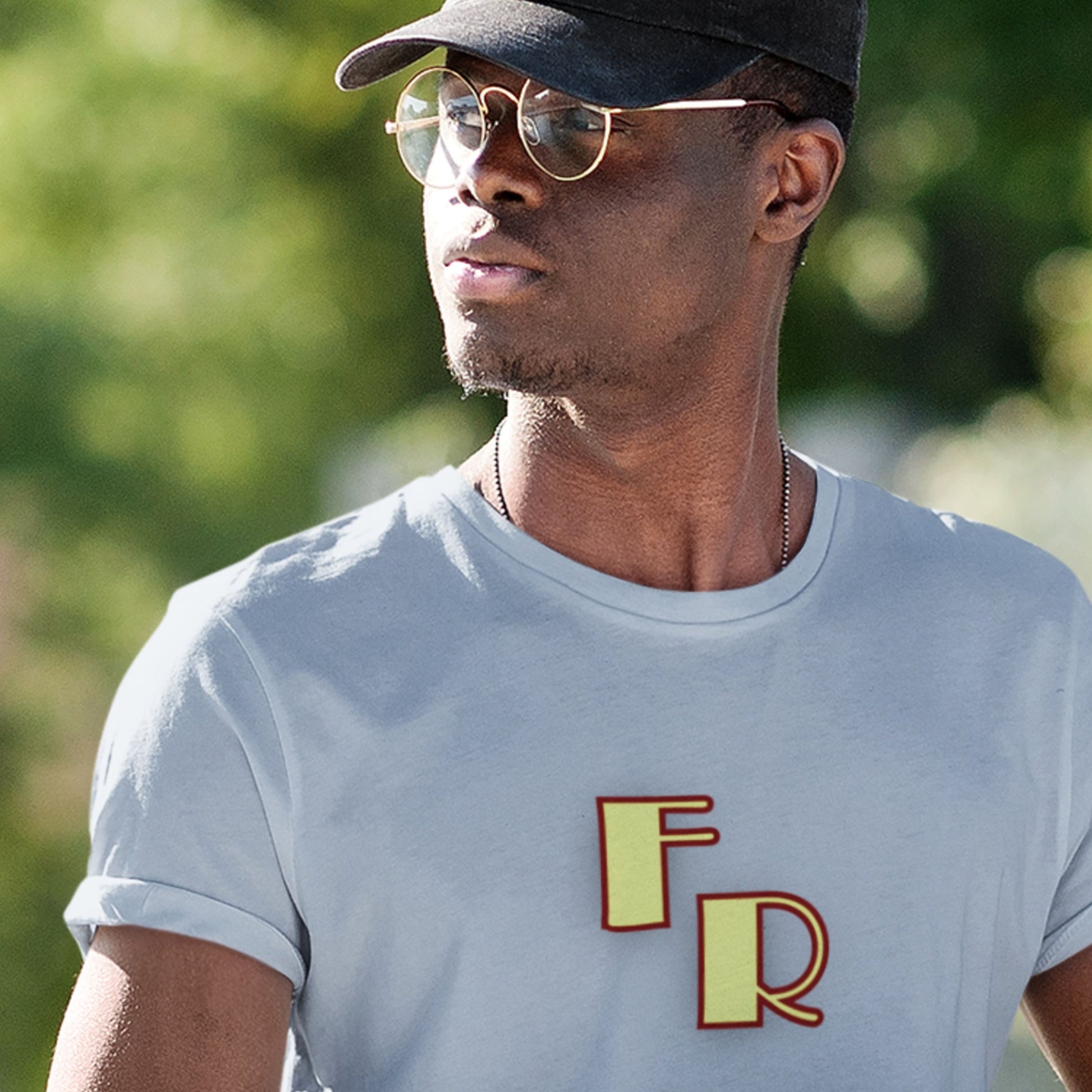Man wearing a light blue t-shirt with 'FR' logo, black cap, and sunglasses outdoors.