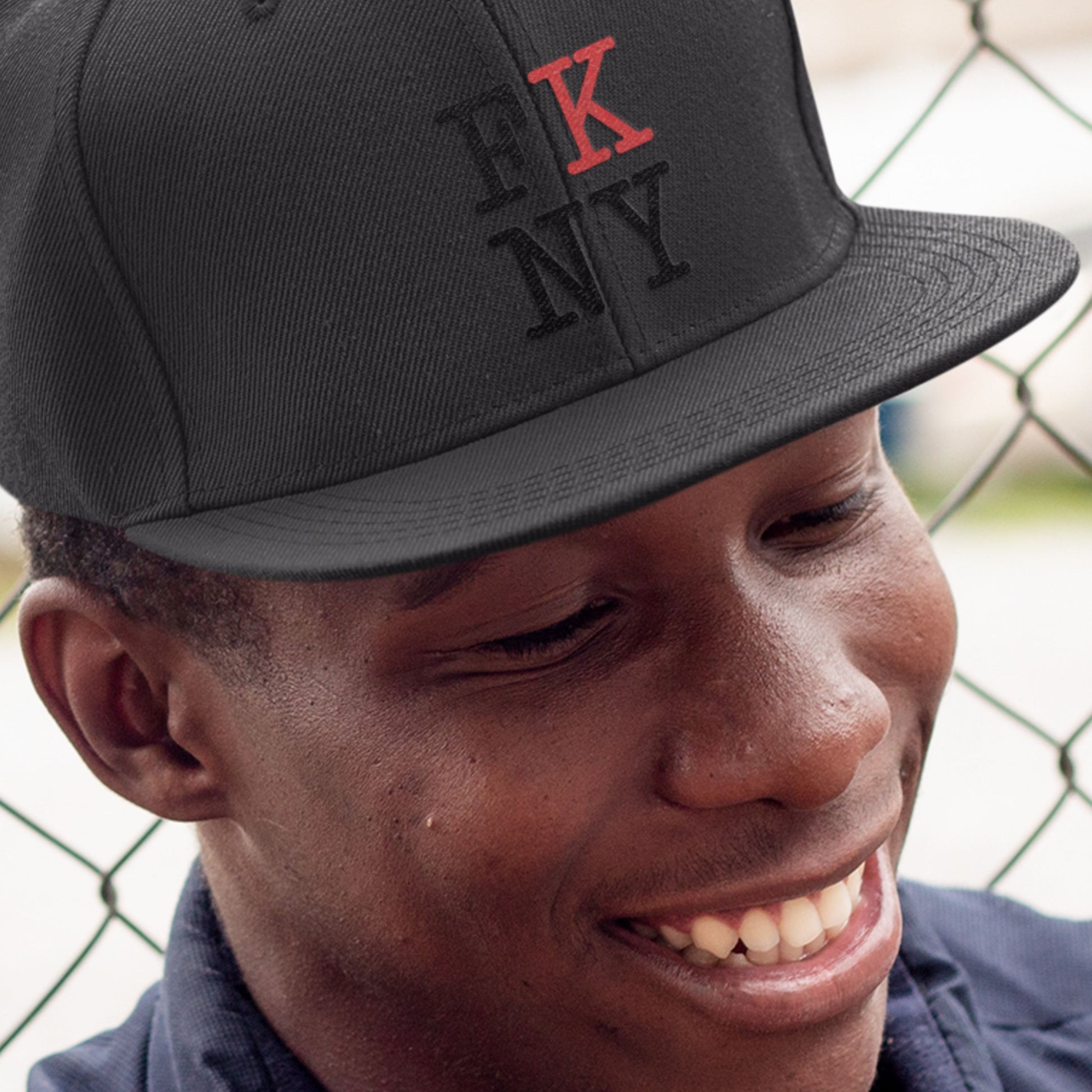 Person wearing a dark grey cap with red and white FK NY logo, smiling outdoors.