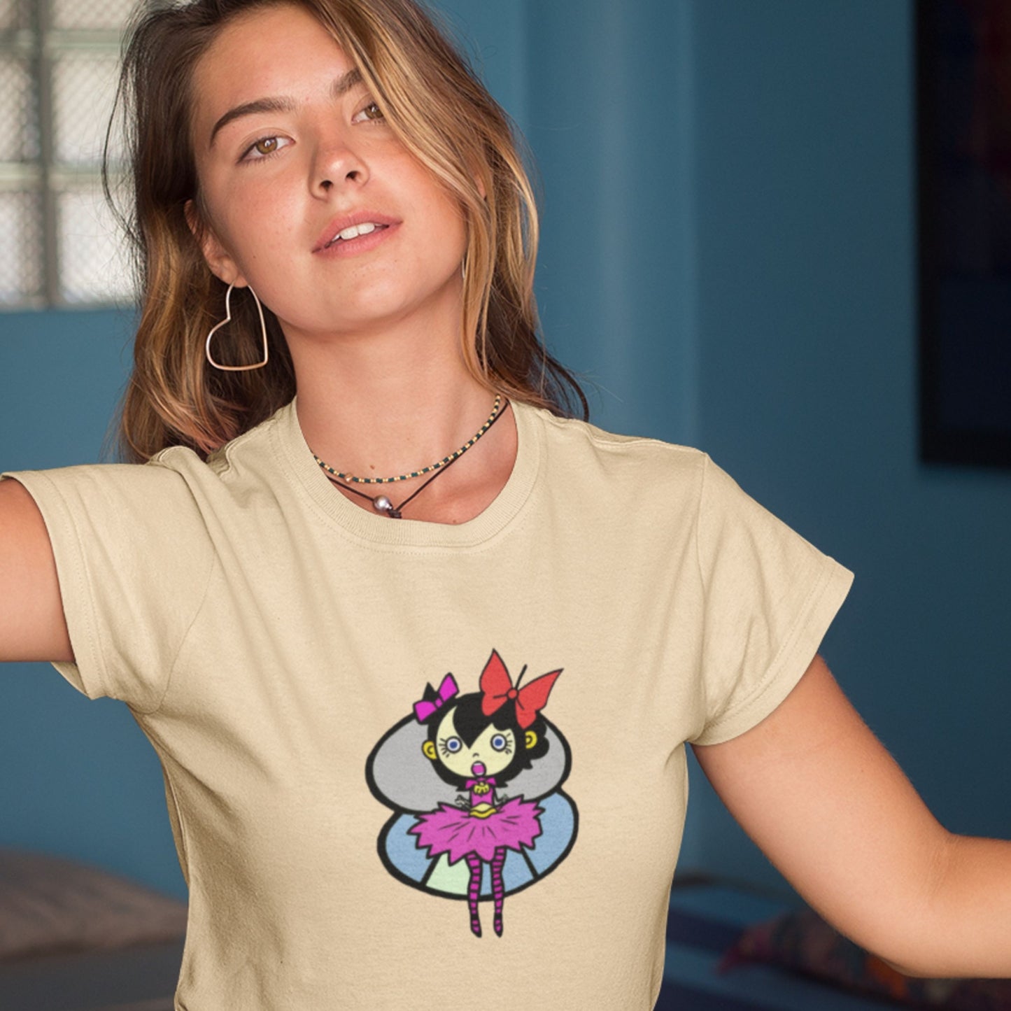 Woman with too-big of earrings wearing a cream colored t-shirt with a fun, colorful cartoon pixie illustration indoors.