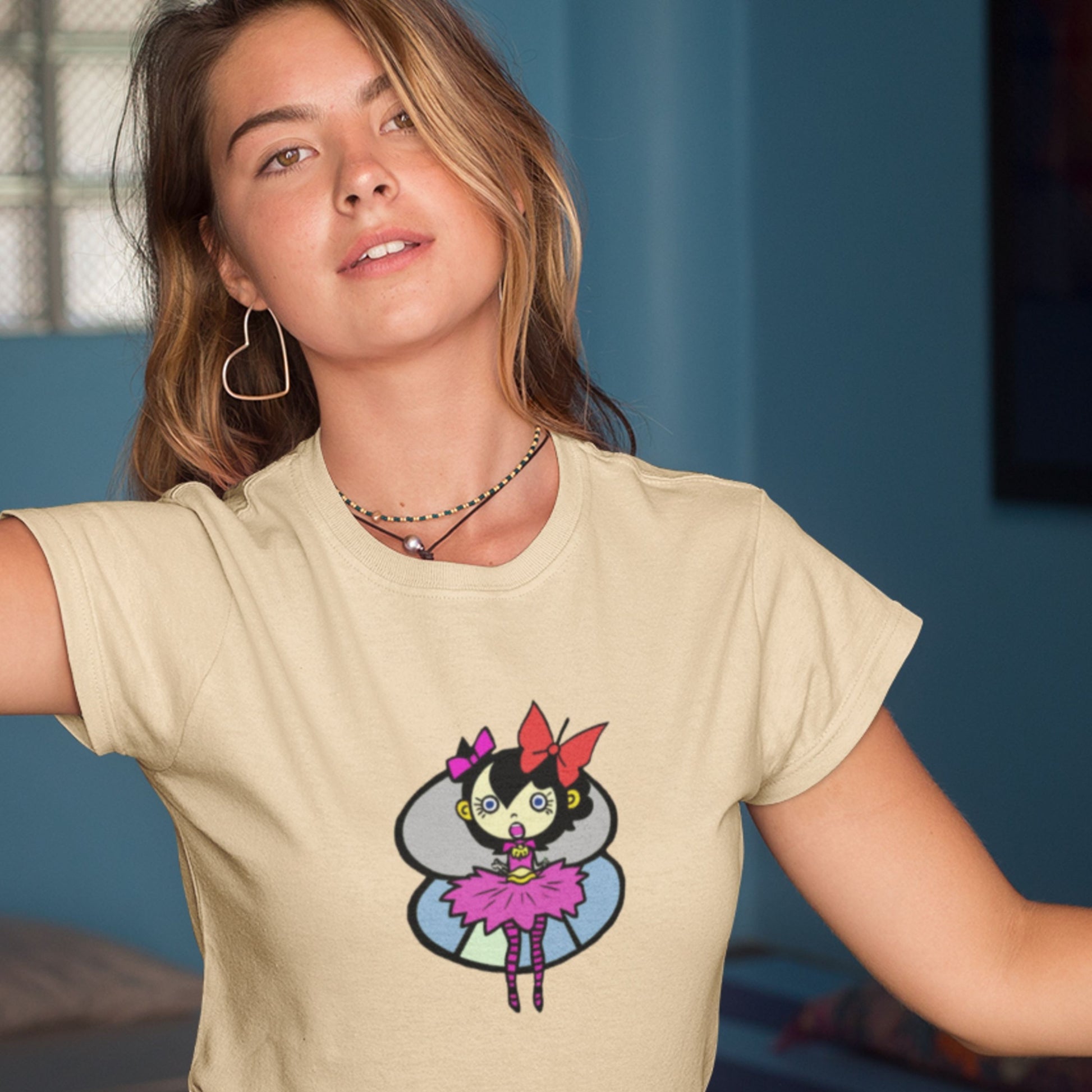 Woman with too-big of earrings wearing a cream colored t-shirt with a fun, colorful cartoon pixie illustration indoors.
