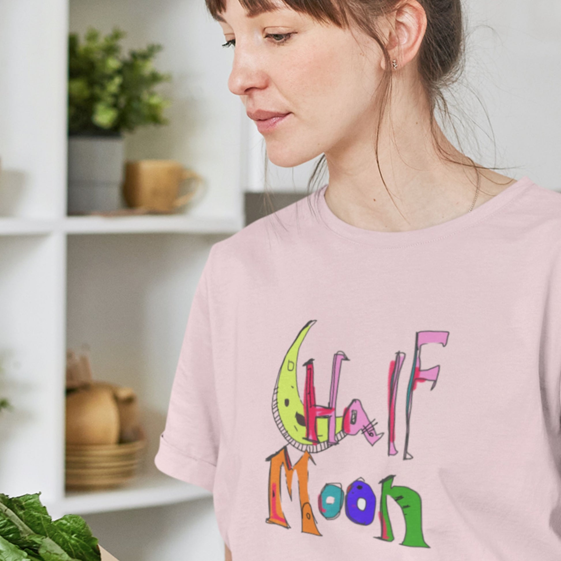 Person wearing a pink t-shirt with colorful text and graphics, standing in a room with shelves and plants.