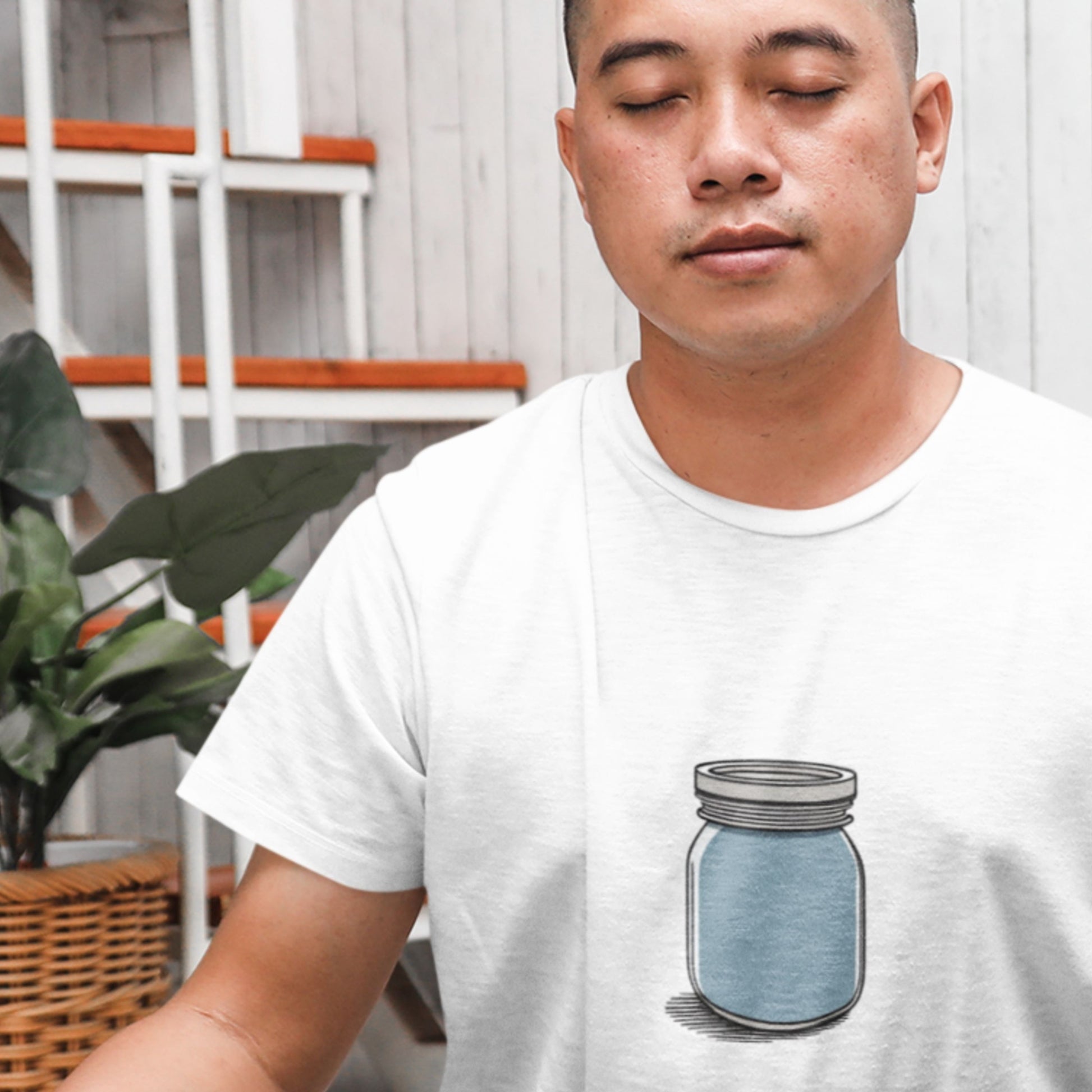 Meditating man wearing a white shirt with a drawing of a blue jar in a living room.