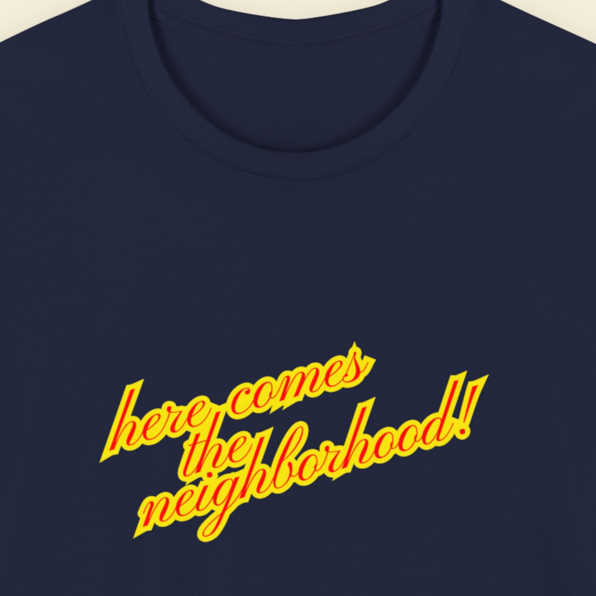 Navy t-shirt with multicolored 'Here Comes the Neighborhood!' text on a beige background.