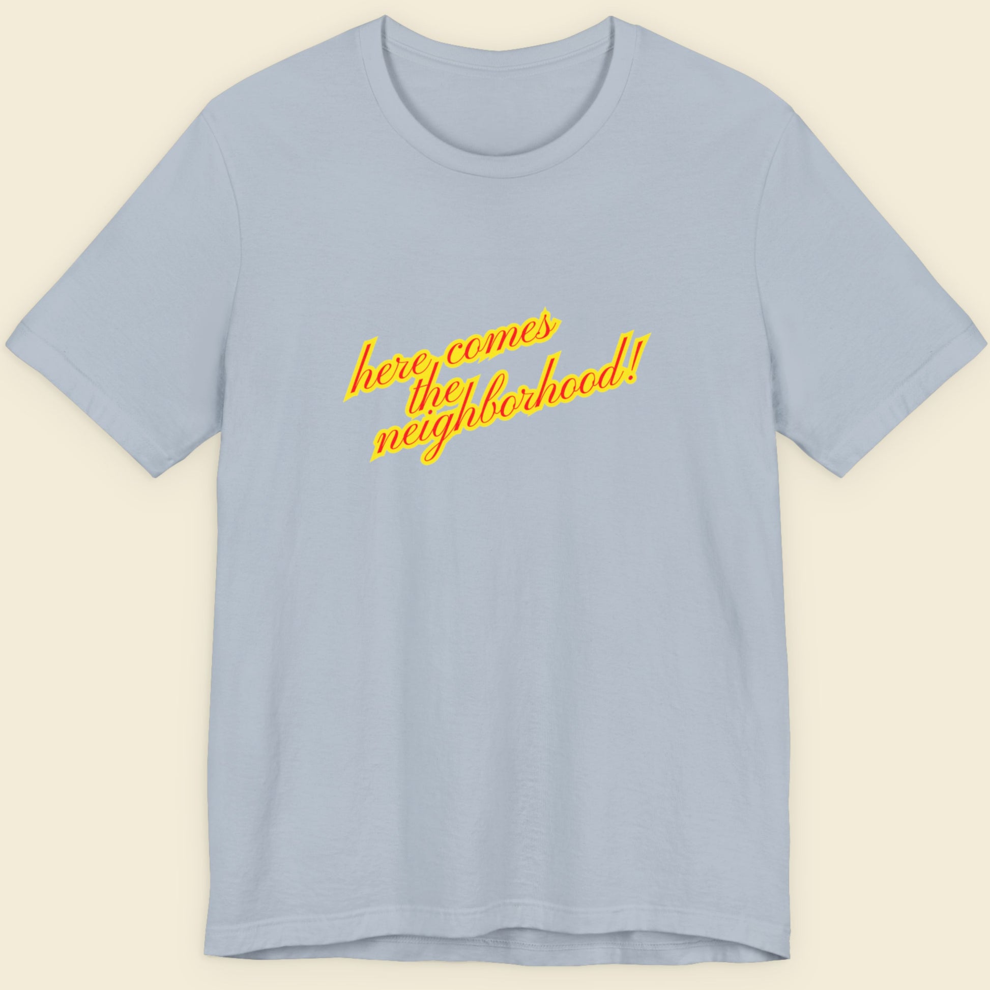 Light blue t-shirt with multicolored 'Here Comes the Neighborhood!' text on a beige background.