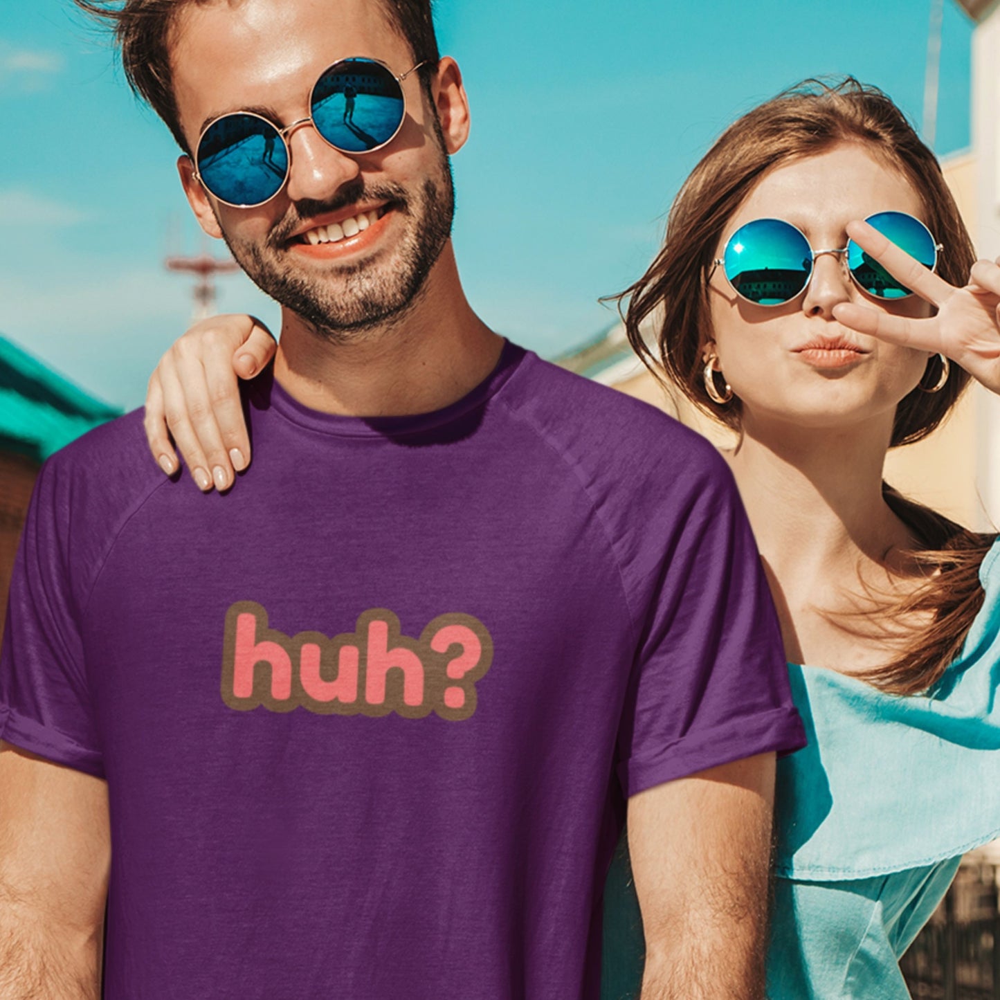 Dude wearing a purple t-shirt with multicolored 'huh?' text at a marina with his girl.