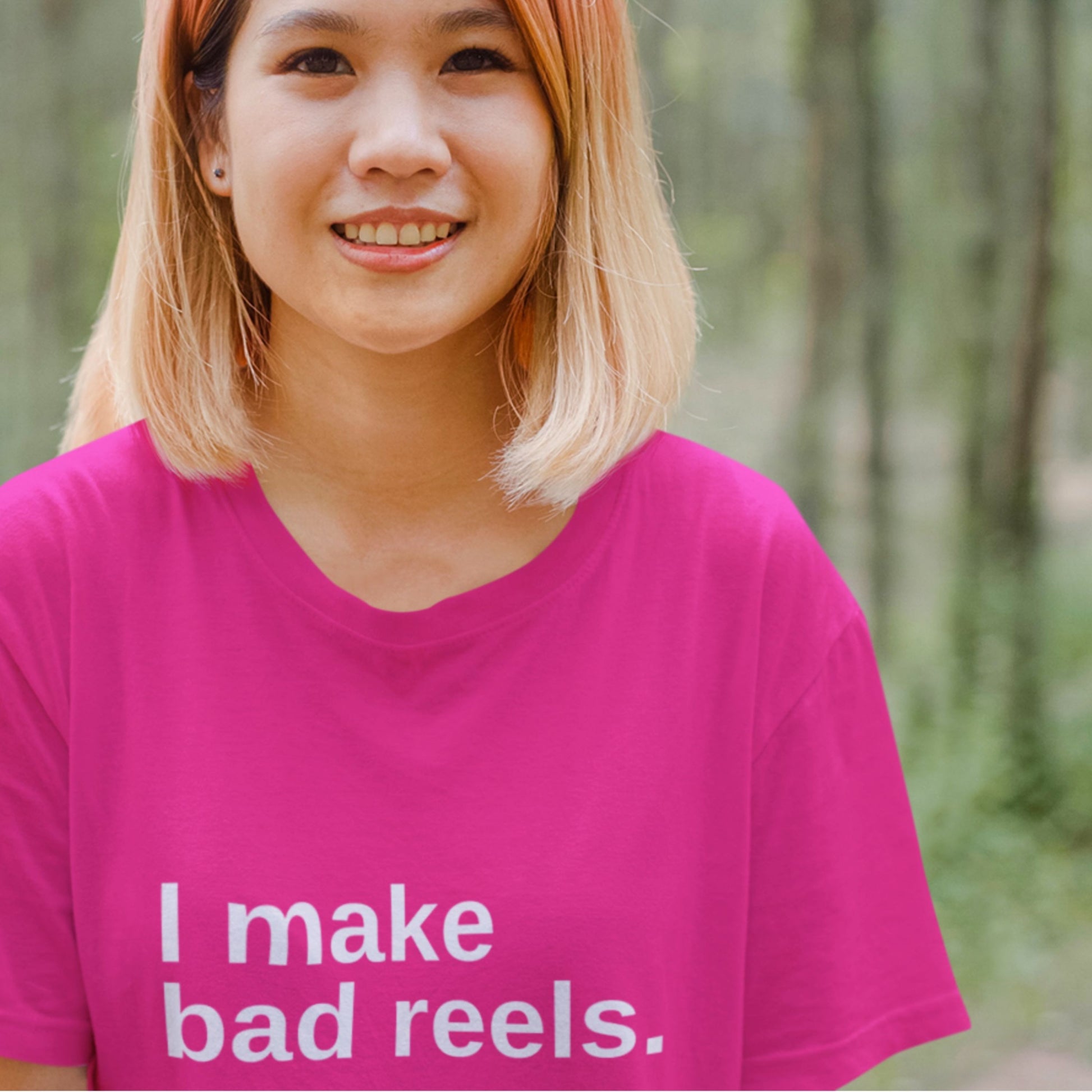 Person wearing a pink shirt with 'I make bad reels' text in a forest setting.