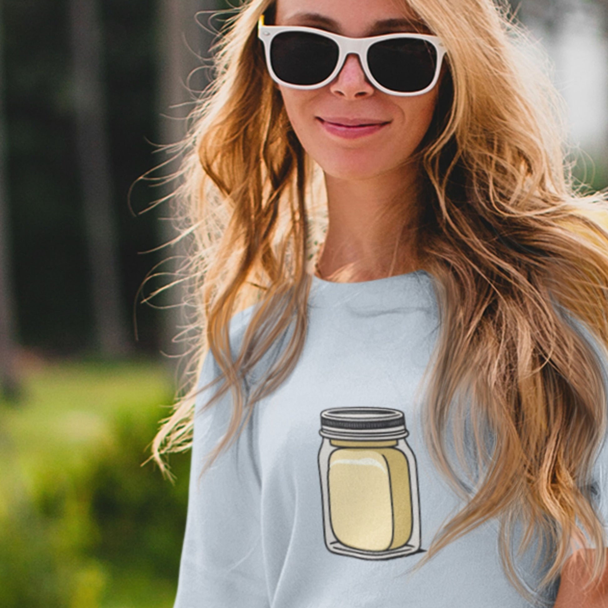 Woman wearing sunglasses and a light blue t-shirt with a jar design outdoors.