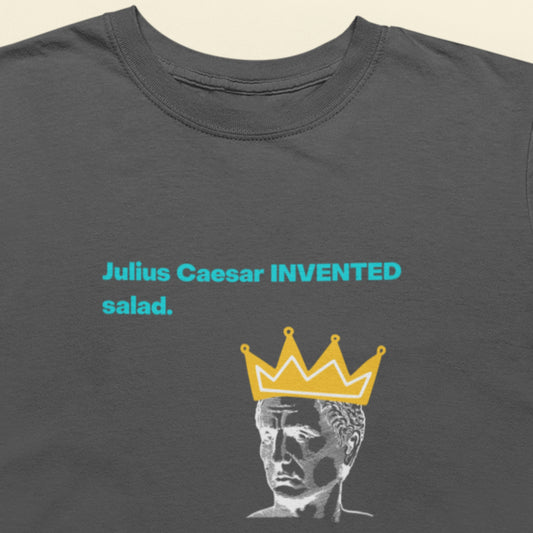 Gray t-shirt with a graphic of a statue wearing a crown and text 'Julius Caesar INVENTED salad' on a beige background.