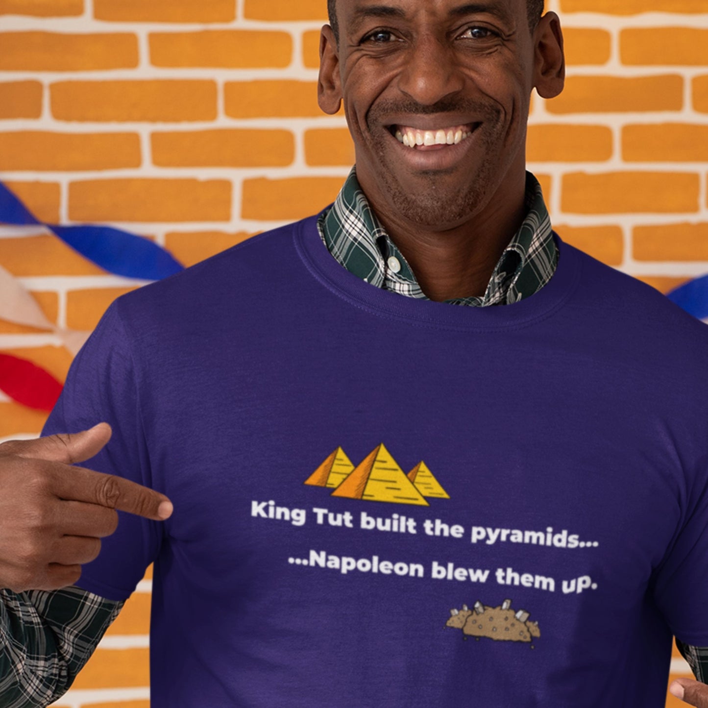 Man wearing a purple t-shirt with a humorous pyramid design and text, pointing at the shirt against a colorful brick wall background.