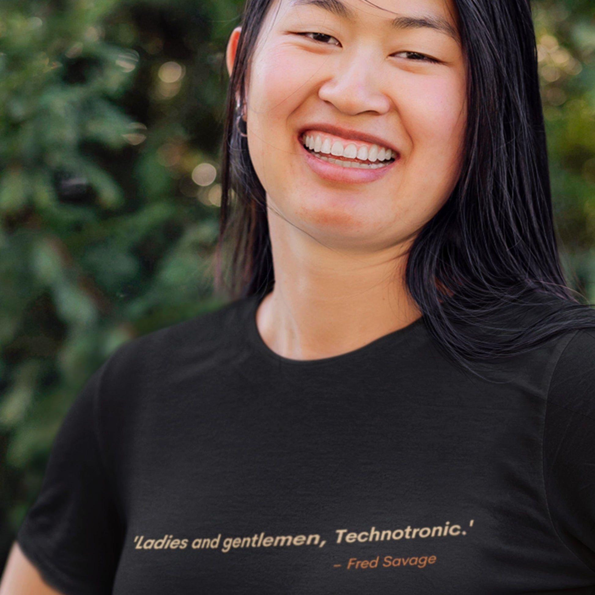 Person wearing a black t-shirt with text, standing outdoors with greenery in the background.