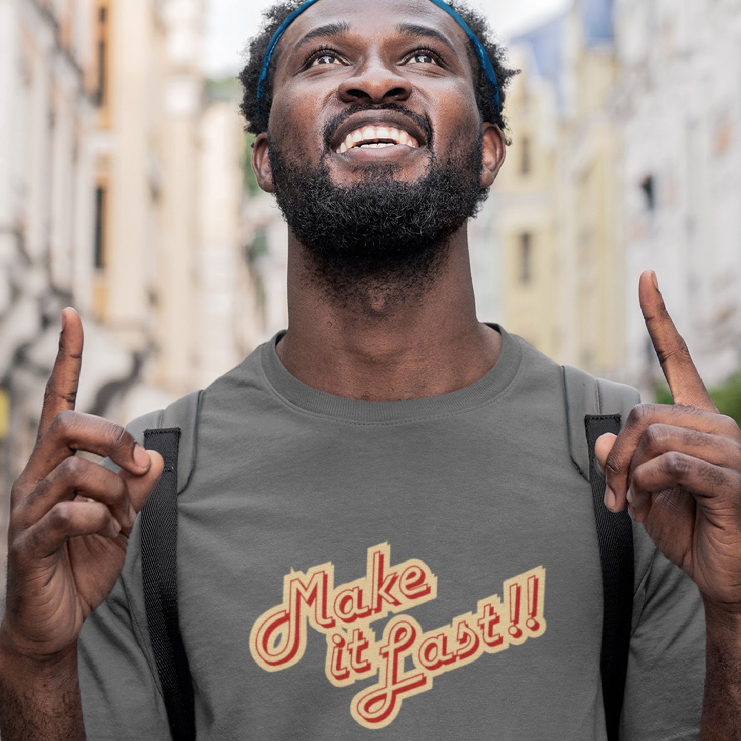 Man wearing a grey t-shirt with 'make it last!!' text in yellow and red in a city.