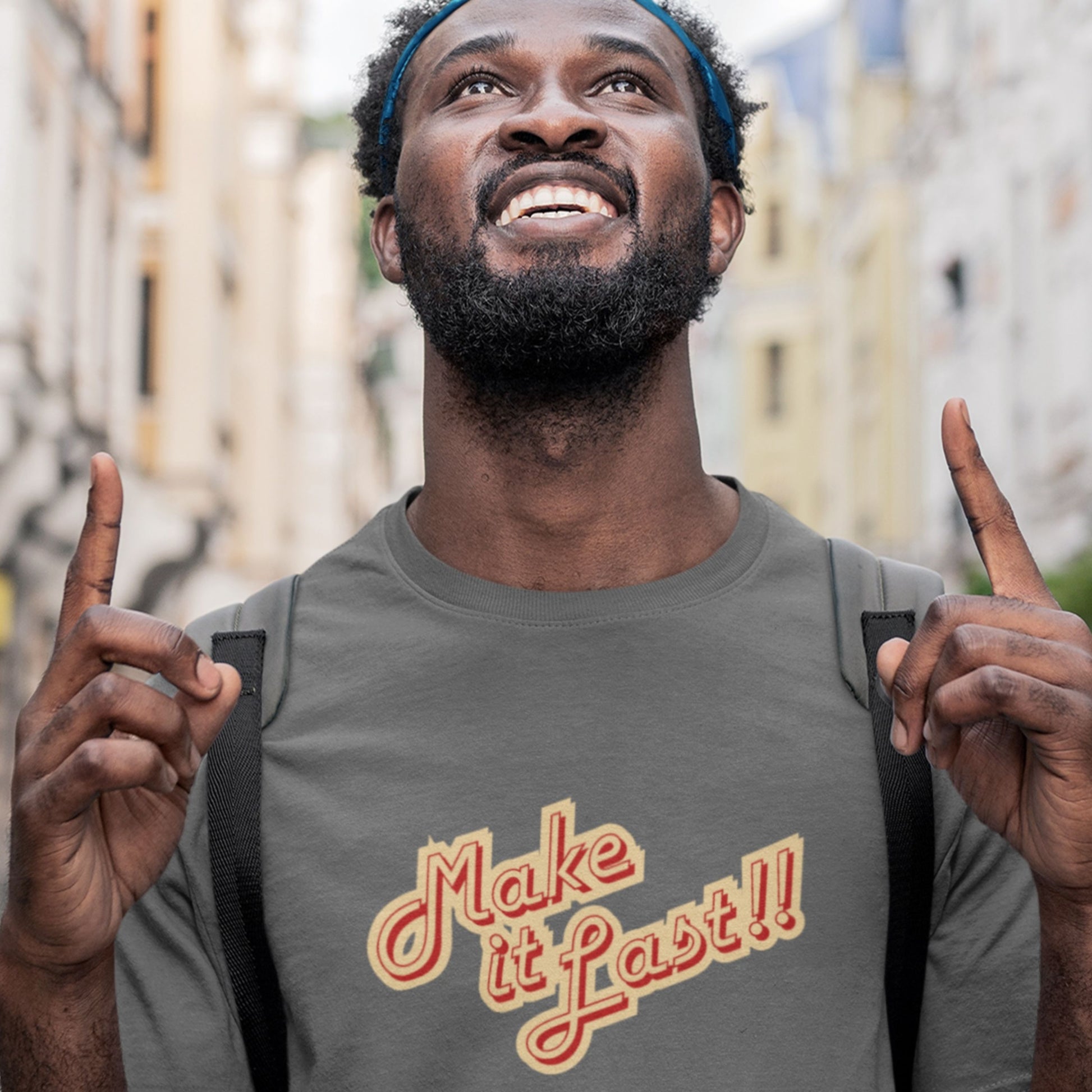 Man wearing a grey t-shirt with 'make it last!!' text in yellow and red in a city.