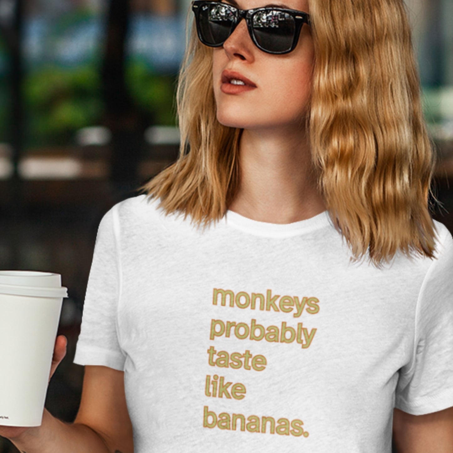 Person wearing a white t-shirt with humorous orange and yellow 'monkeys probably taste like bananas' text, holding a coffee cup.