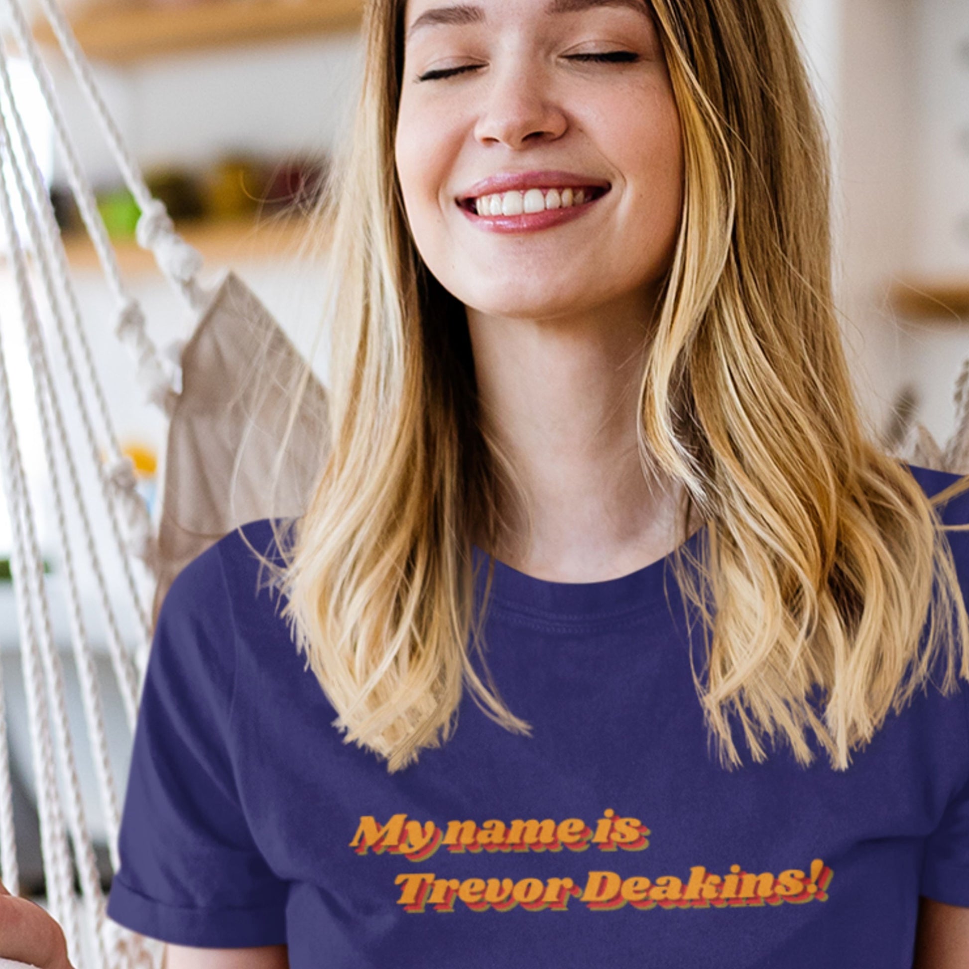 Woman wearing a purple t-shirt with 'My name is Trevor Deakins!' text, smiling.