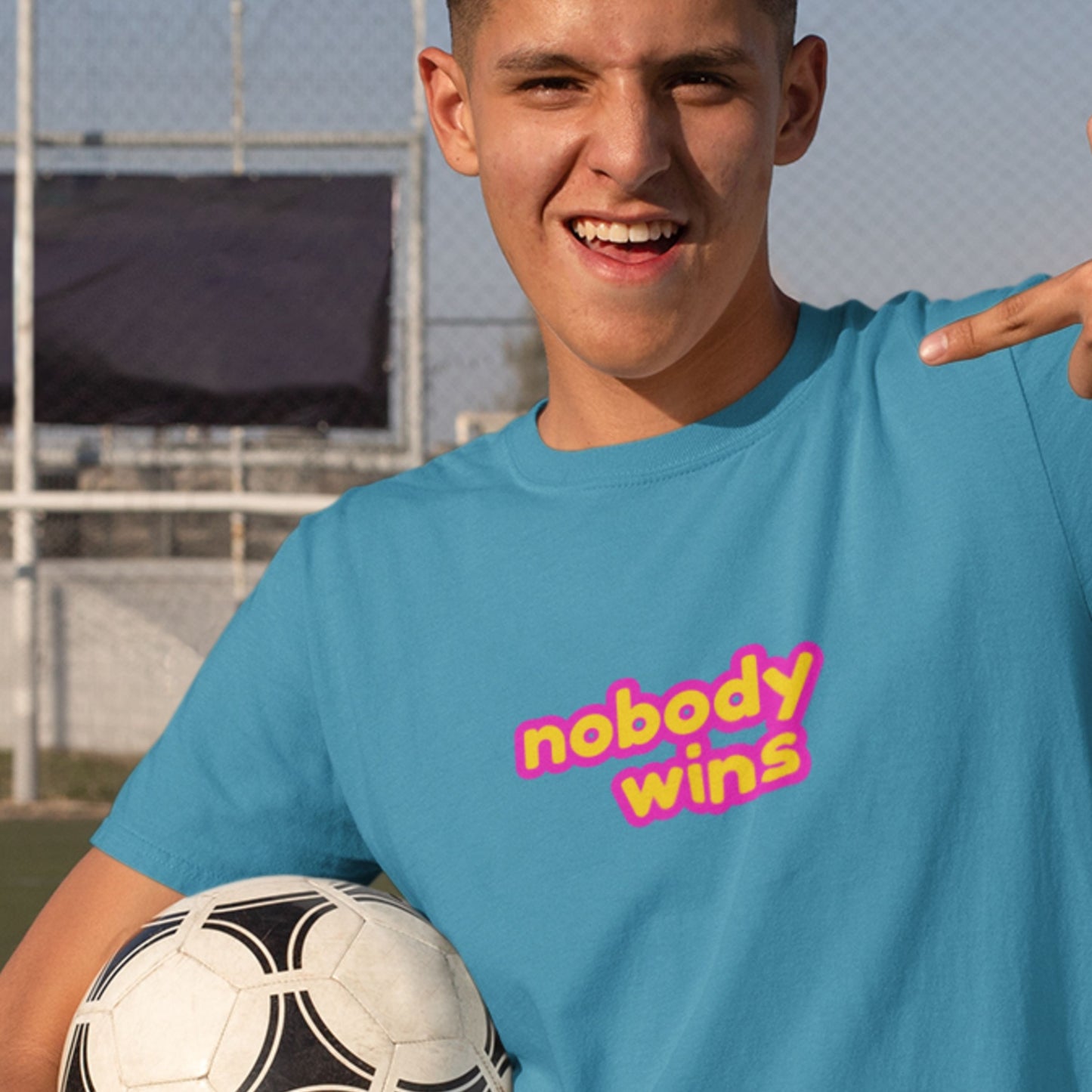 Soccer boy wearing a blue tee with multicolored 'nobody wins' text on a soccer pitch.