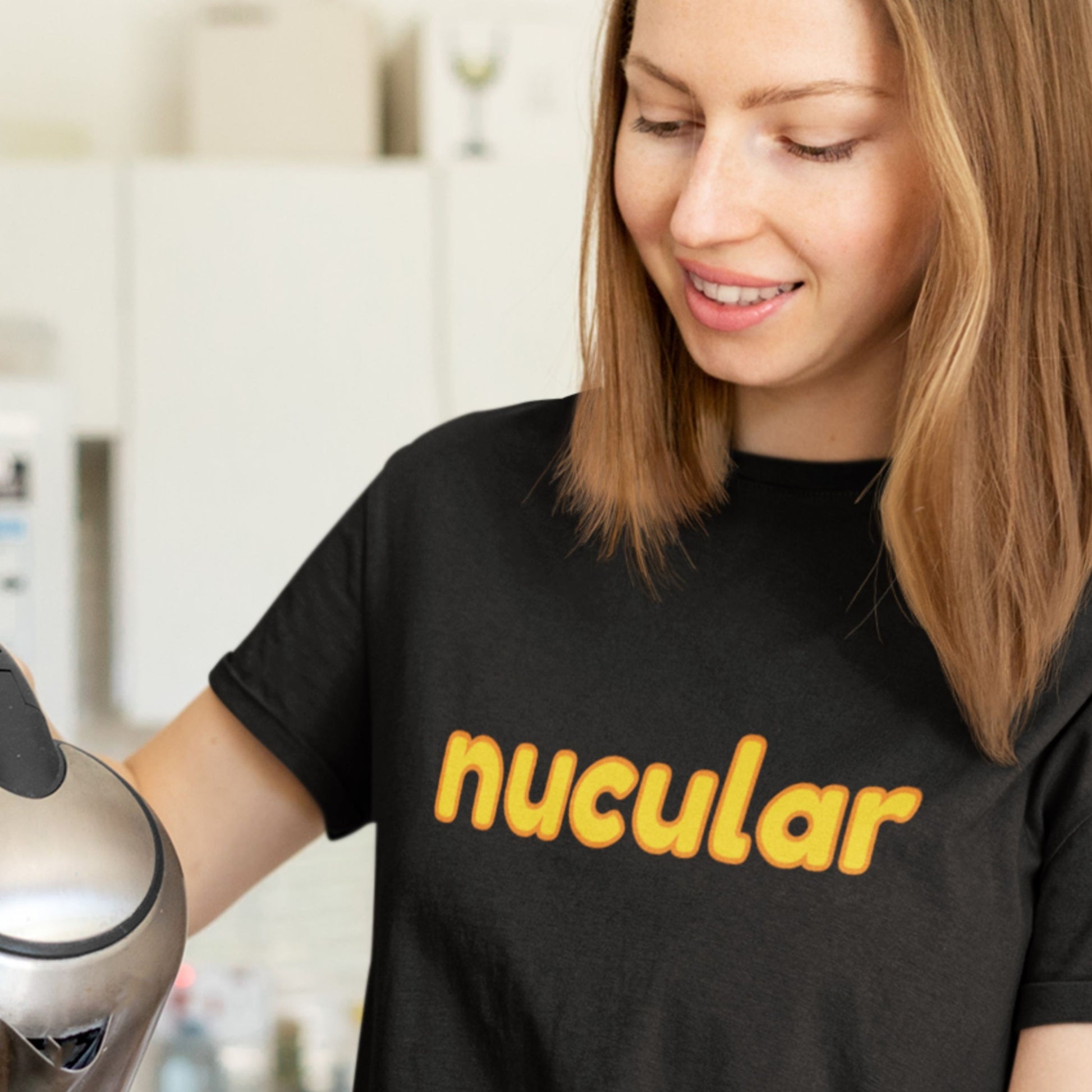 Woman wearing a black t-shirt with 'nucular' printed in yellow, holding a coffee maker.