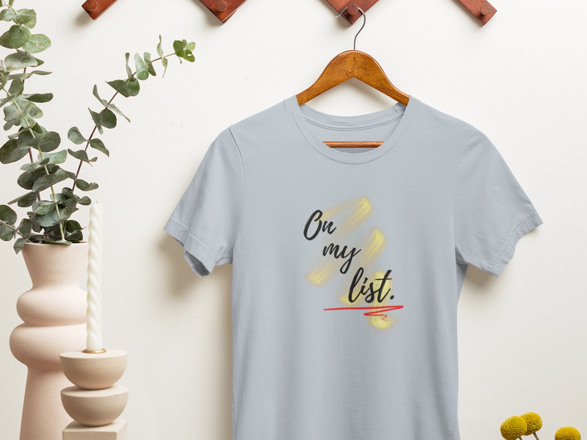 Light blue t-shirt with 'On my list' text on a wooden hanger against a white wall.