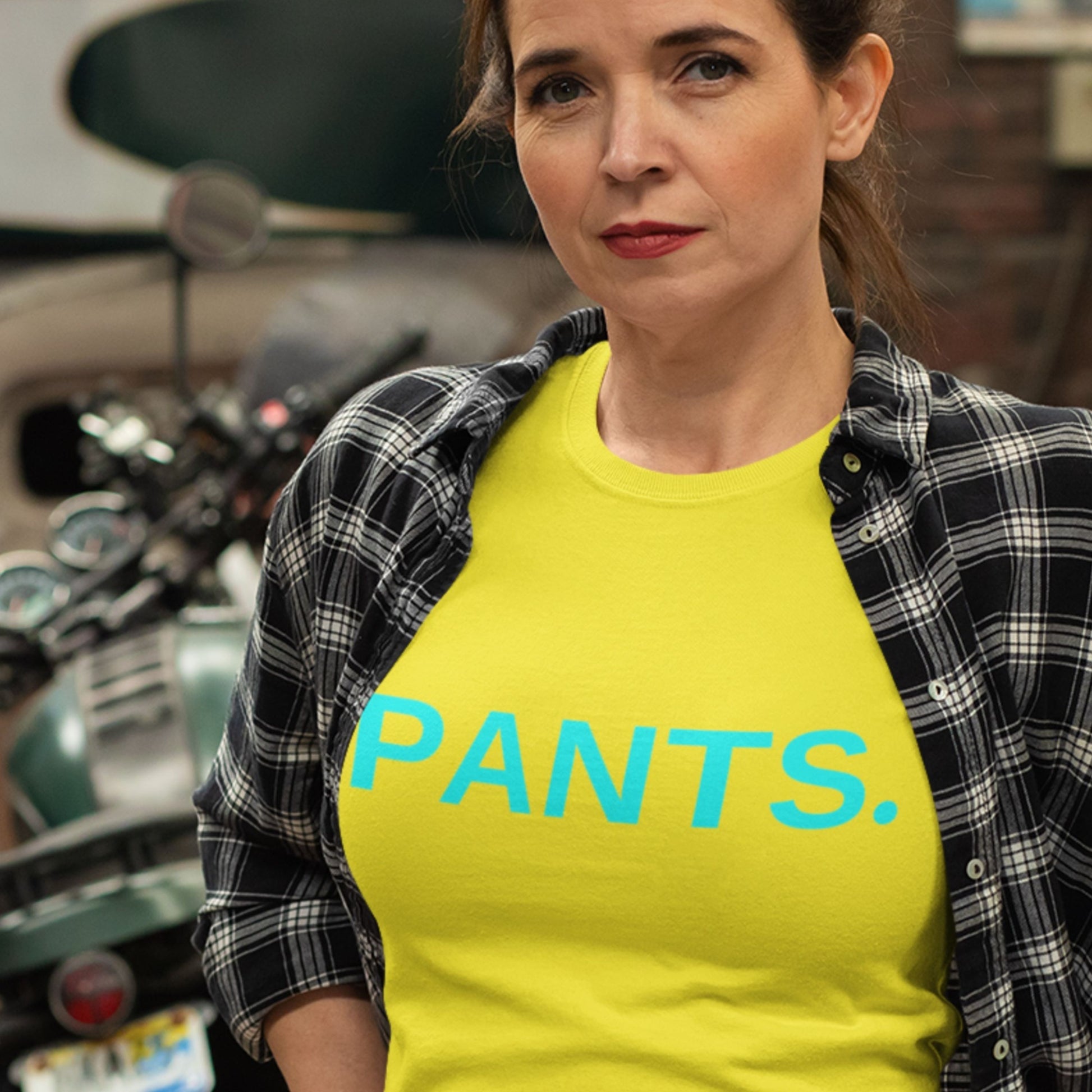 Woman wearing a yellow 'PANTS.' shirt with a plaid jacket, standing in front of a motorcycle.