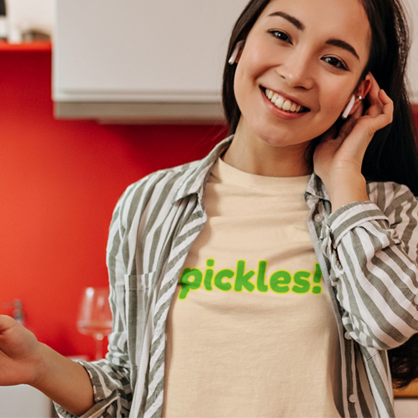 Woman smiles while wearing a cream colored t-shirt with humorous colorful pickles! text in a kitchen.