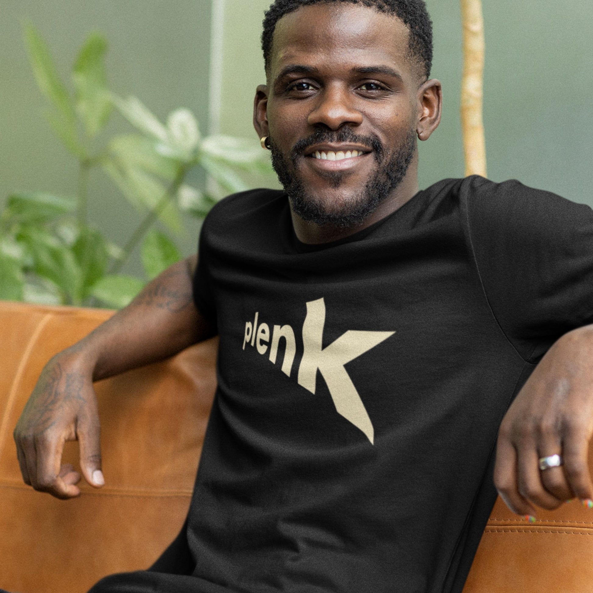 Man wearing a black t-shirt with 'plenK' logo sitting on a brown leather couch.