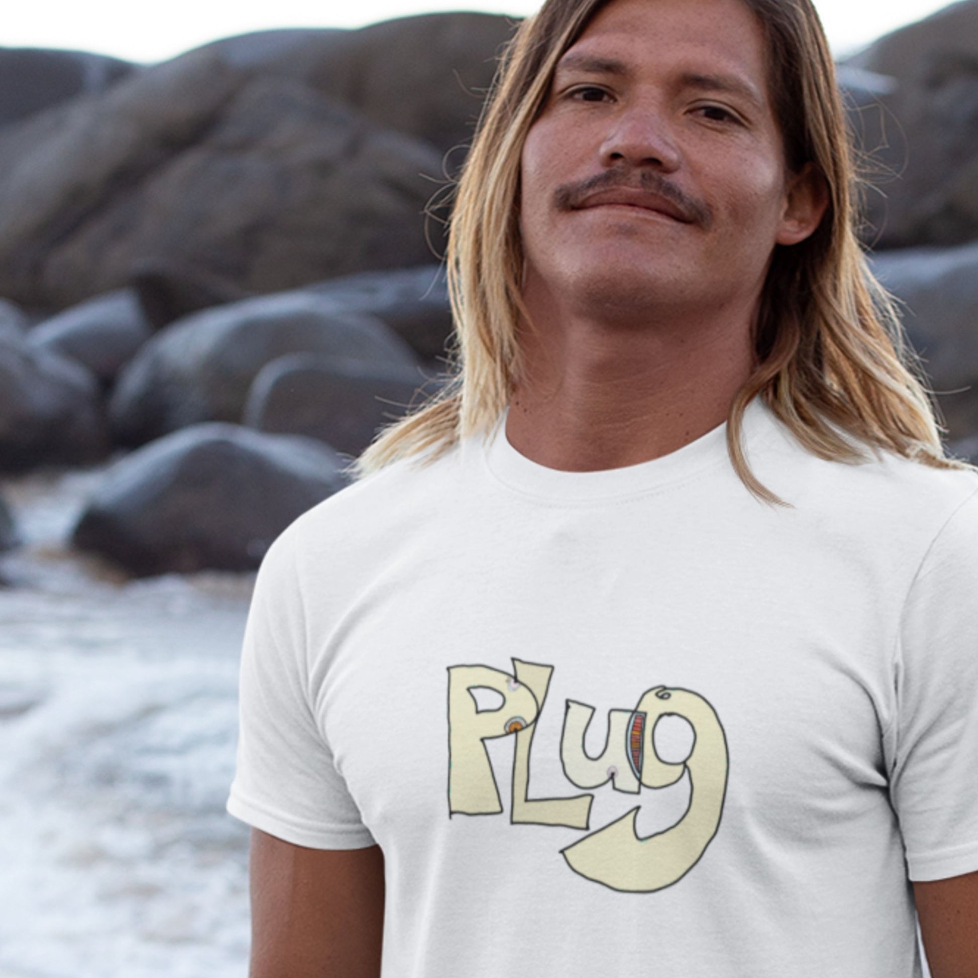 Man wearing a white t-shirt with 'Plug' logo on a rocky beach