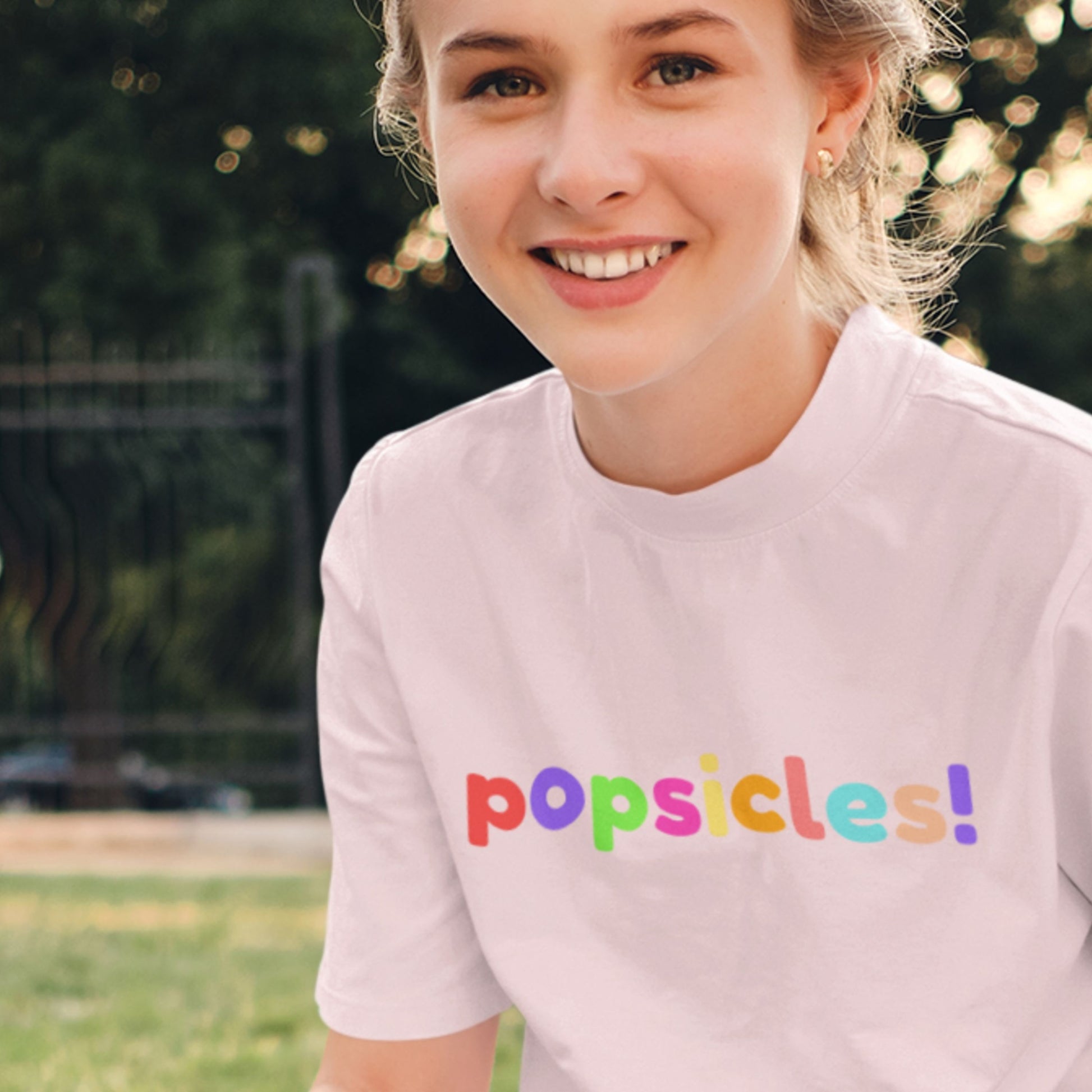 Person wearing a light pink t-shirt with 'popsicles!' in colorful letters outdoors.