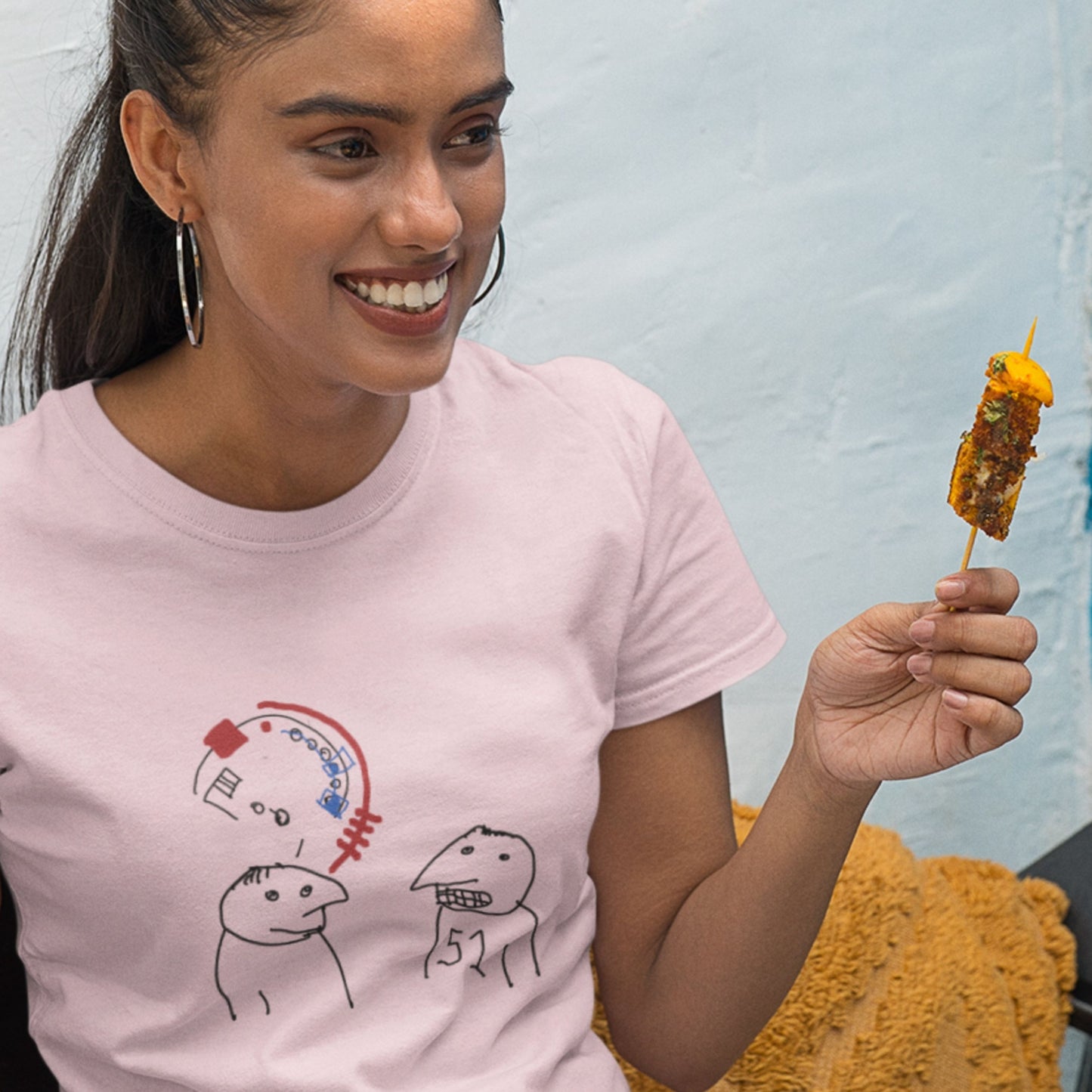 Woman eating some food while wearing a pink t-shirt with a colorful abstract drawing indoors.